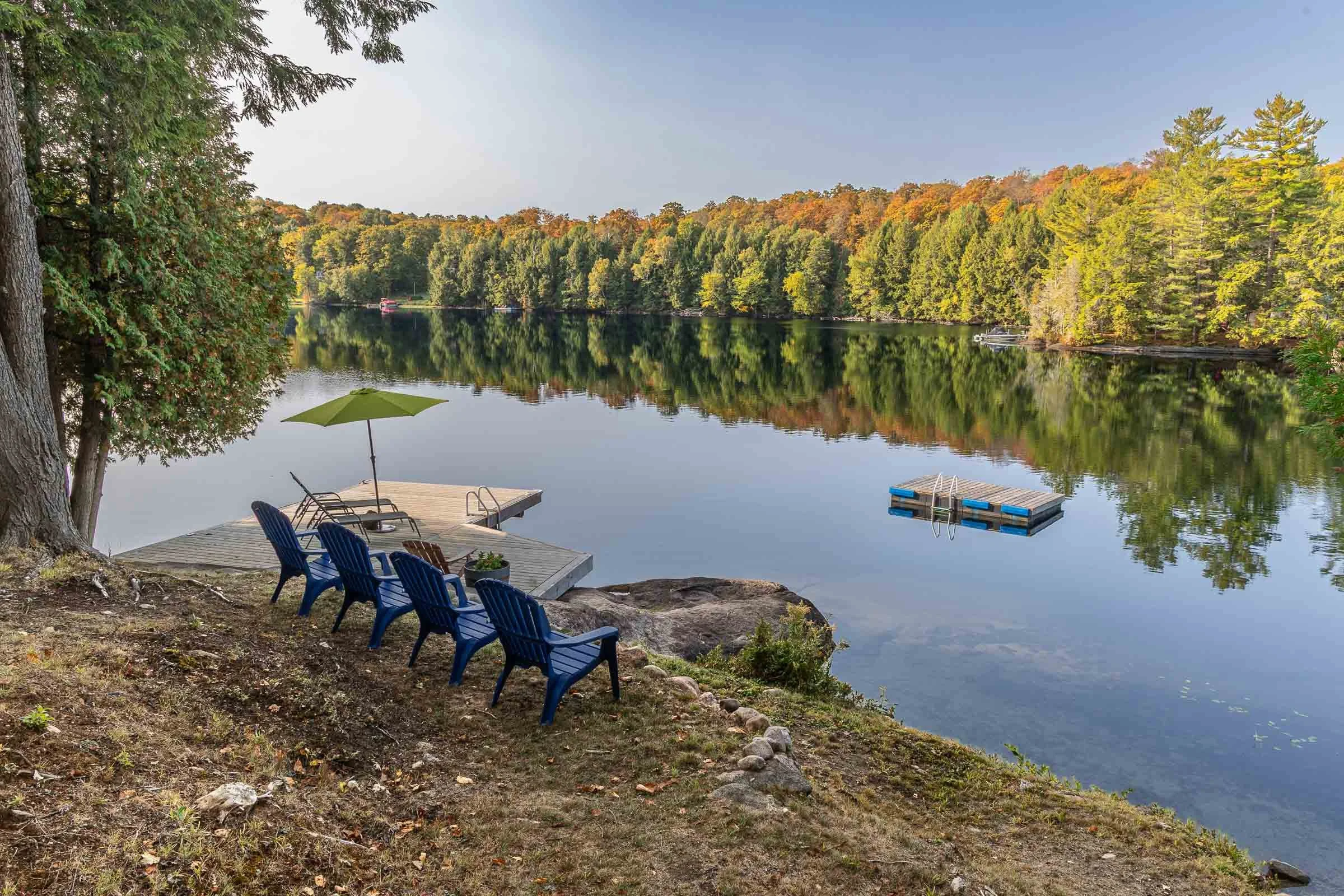 A peaceful lakeside scene with a dock, blue chairs, a green umbrella, and a reflection of fall foliage in the water.