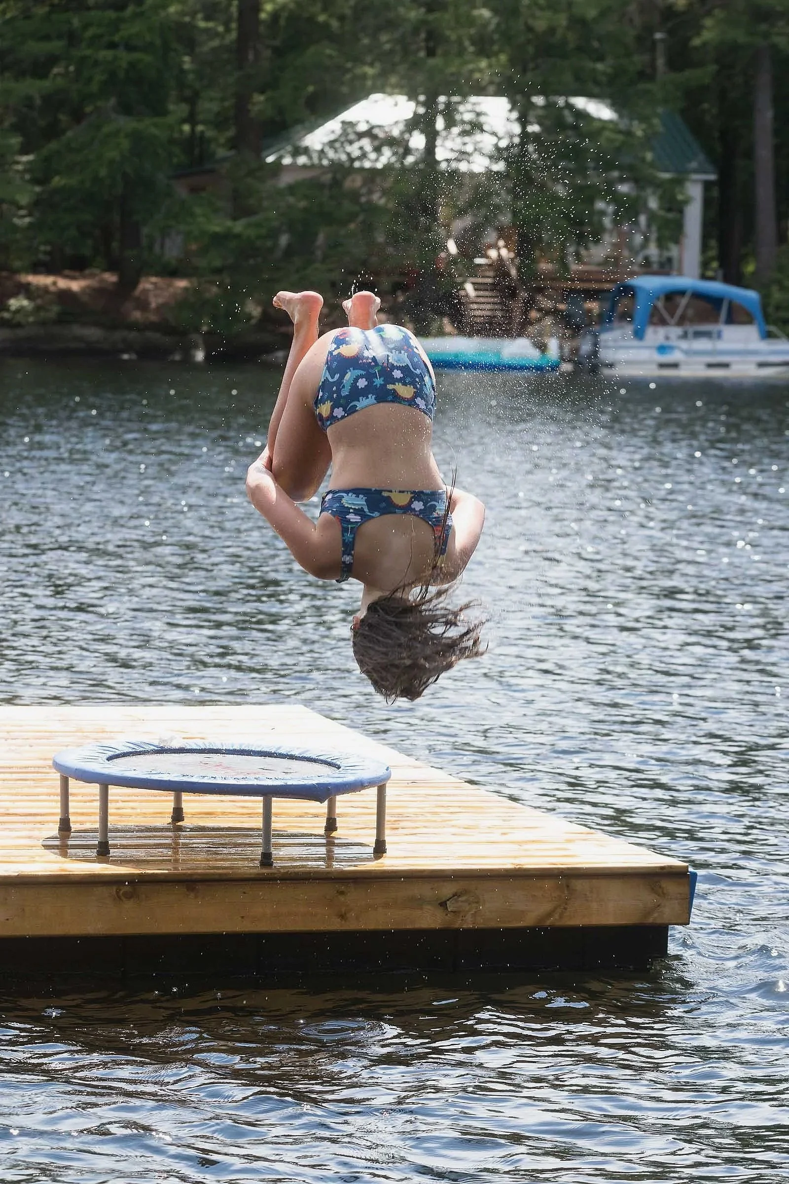 Child flipping into the water off a dock, wearing a patterned swimsuit, with a small trampoline on the dock and a background of trees and boats on a lake.
