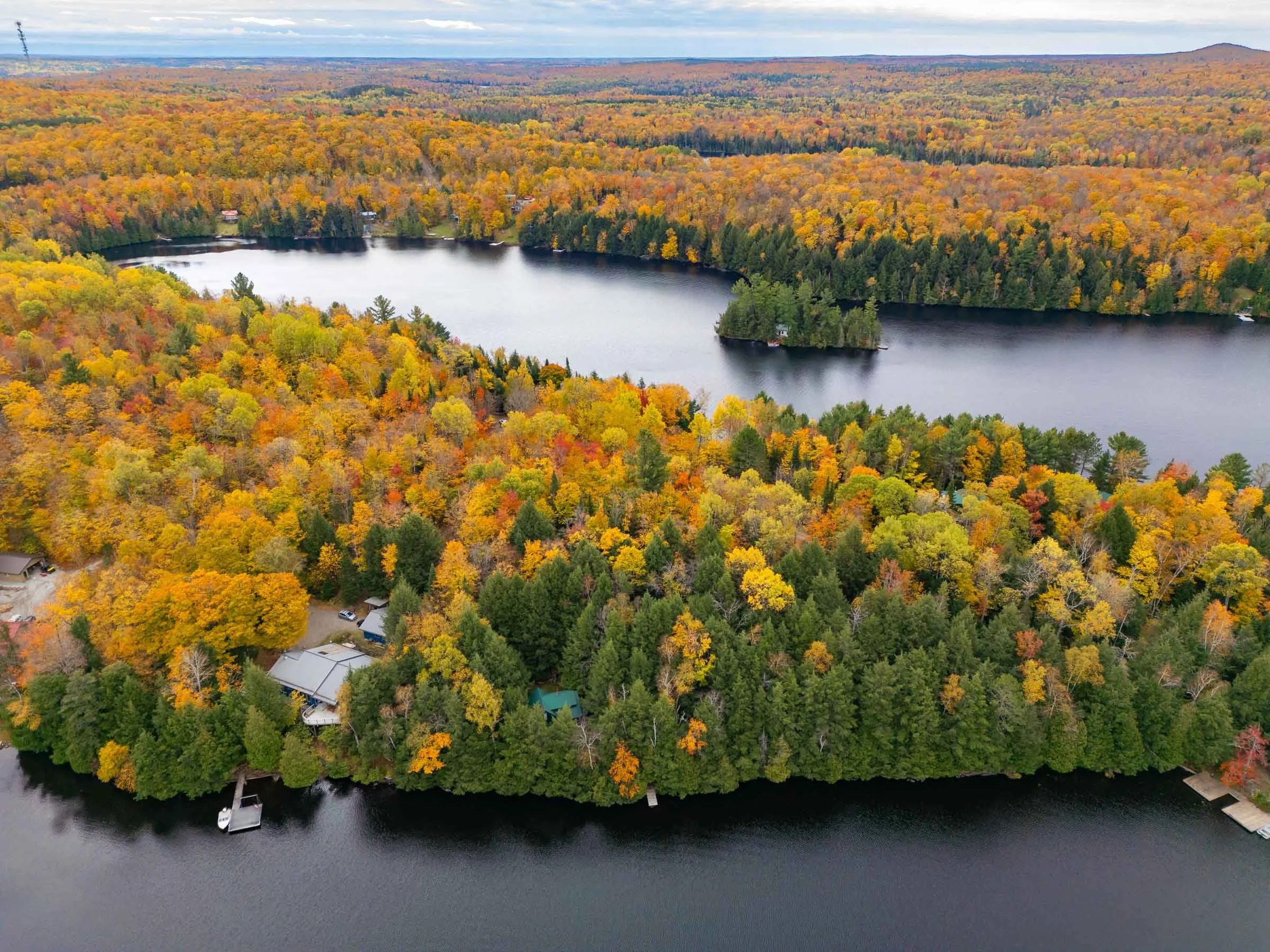 Aerial view of a lake surrounded by dense autumn-colored forest with houses and docks along the shoreline.
