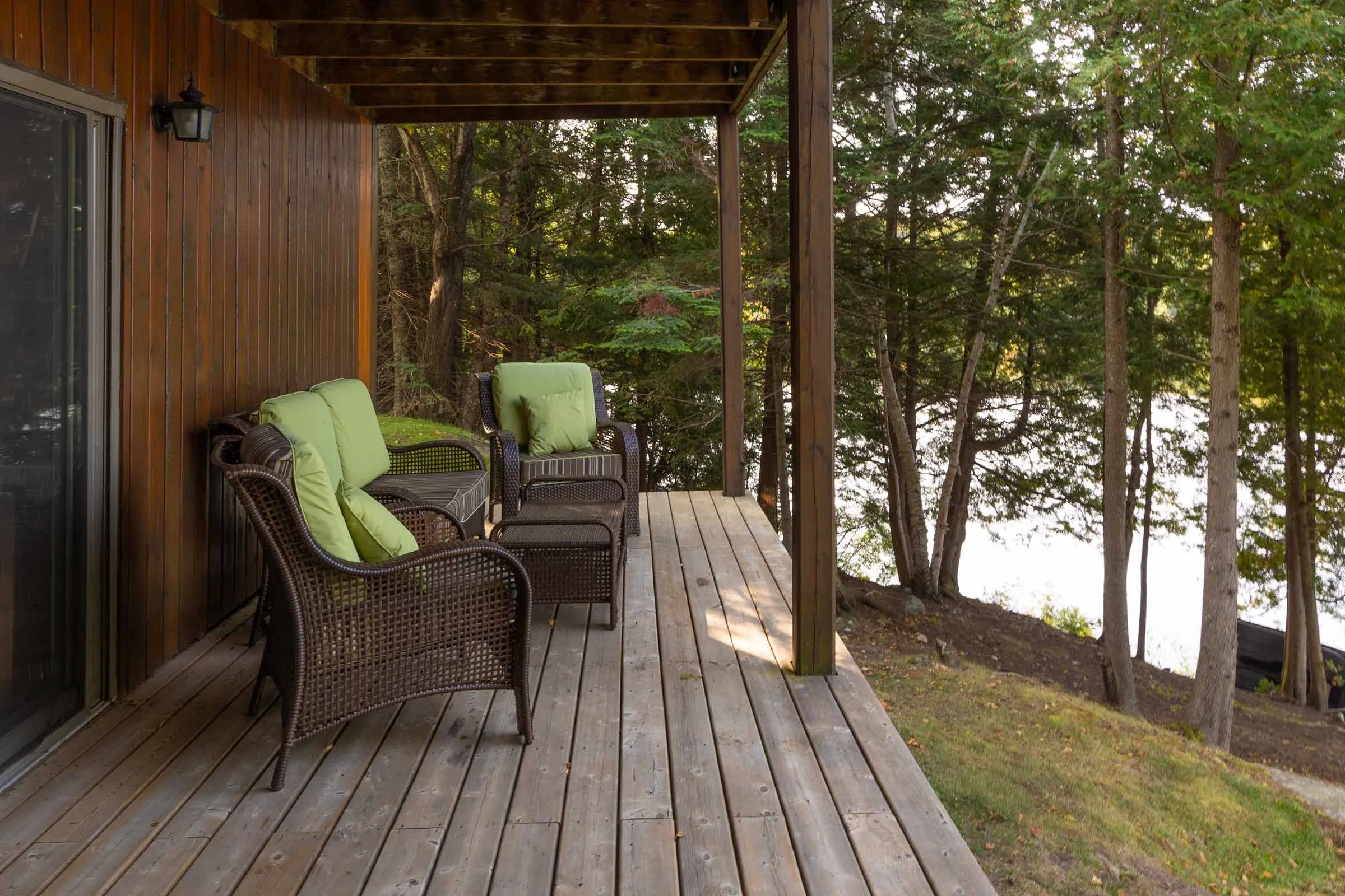A wooden balcony with outdoor wicker chairs and lime green cushions overlooking a forested area with tall trees.