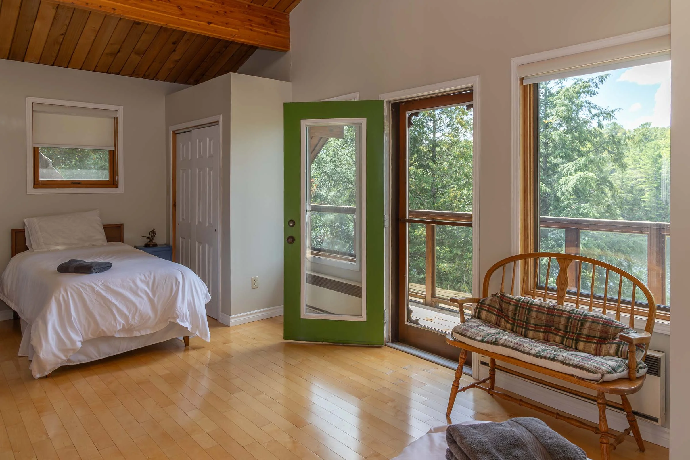 A bedroom with a bed, a window, and a wooden rocking chair with a plaid cushion near the window. The room has hardwood floors, a green accent wall, and large windows showing trees outside.