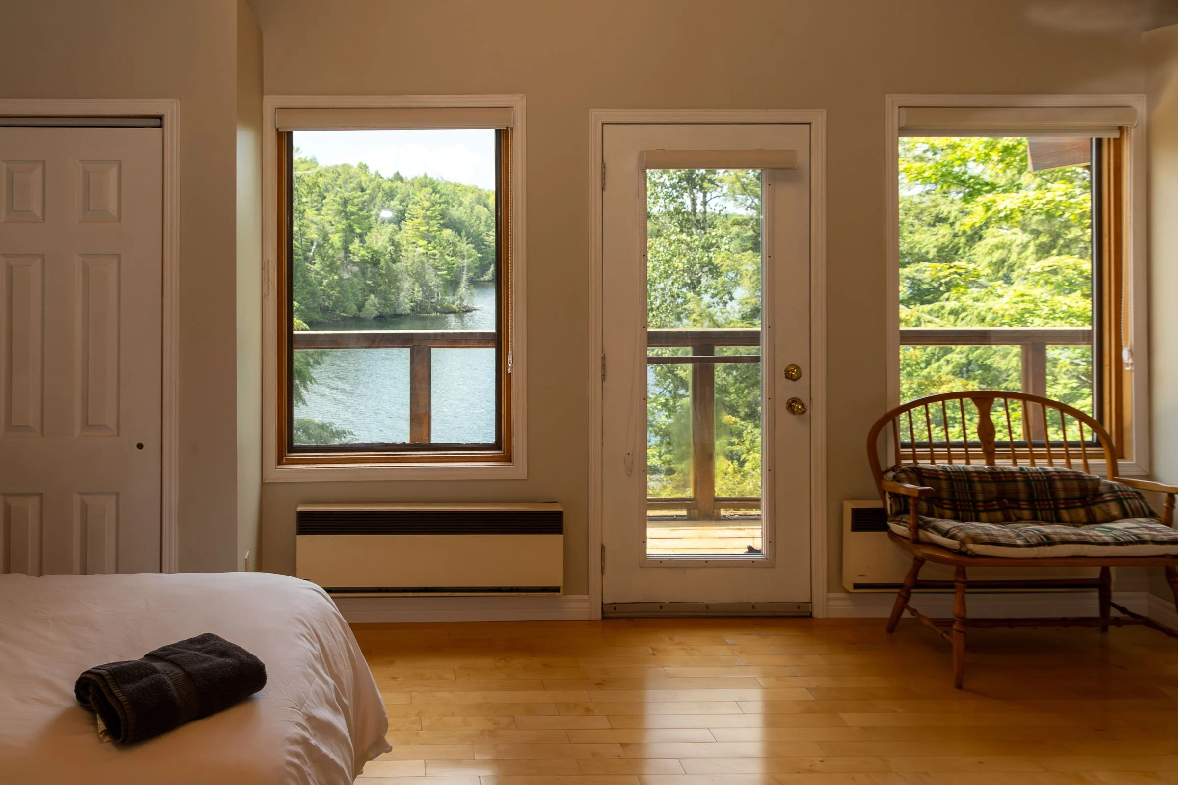 A bedroom interior with three large windows showing a lake and trees outside. There is a bed with folded dark towels at the foot, and a wooden chair near the right window. The door to a balcony is open, overlooking the greenery and water.