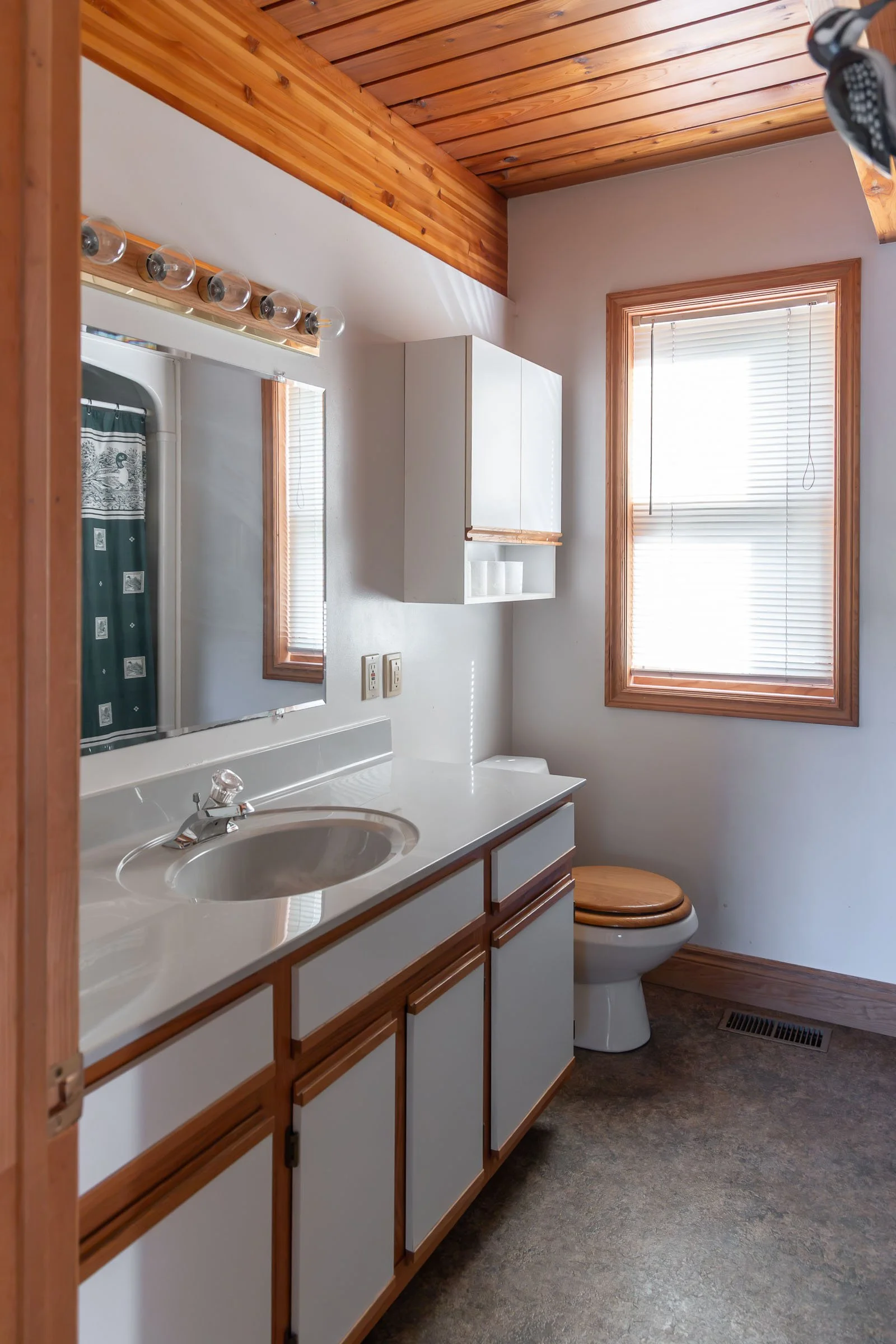 A small bathroom with a wooden ceiling and trim, white walls, a window with blinds, white bathroom cabinet, mirror, sink, and a toilet with a wooden seat. A shower with a green curtain is visible in the mirror, and the bathroom floor has a vent.