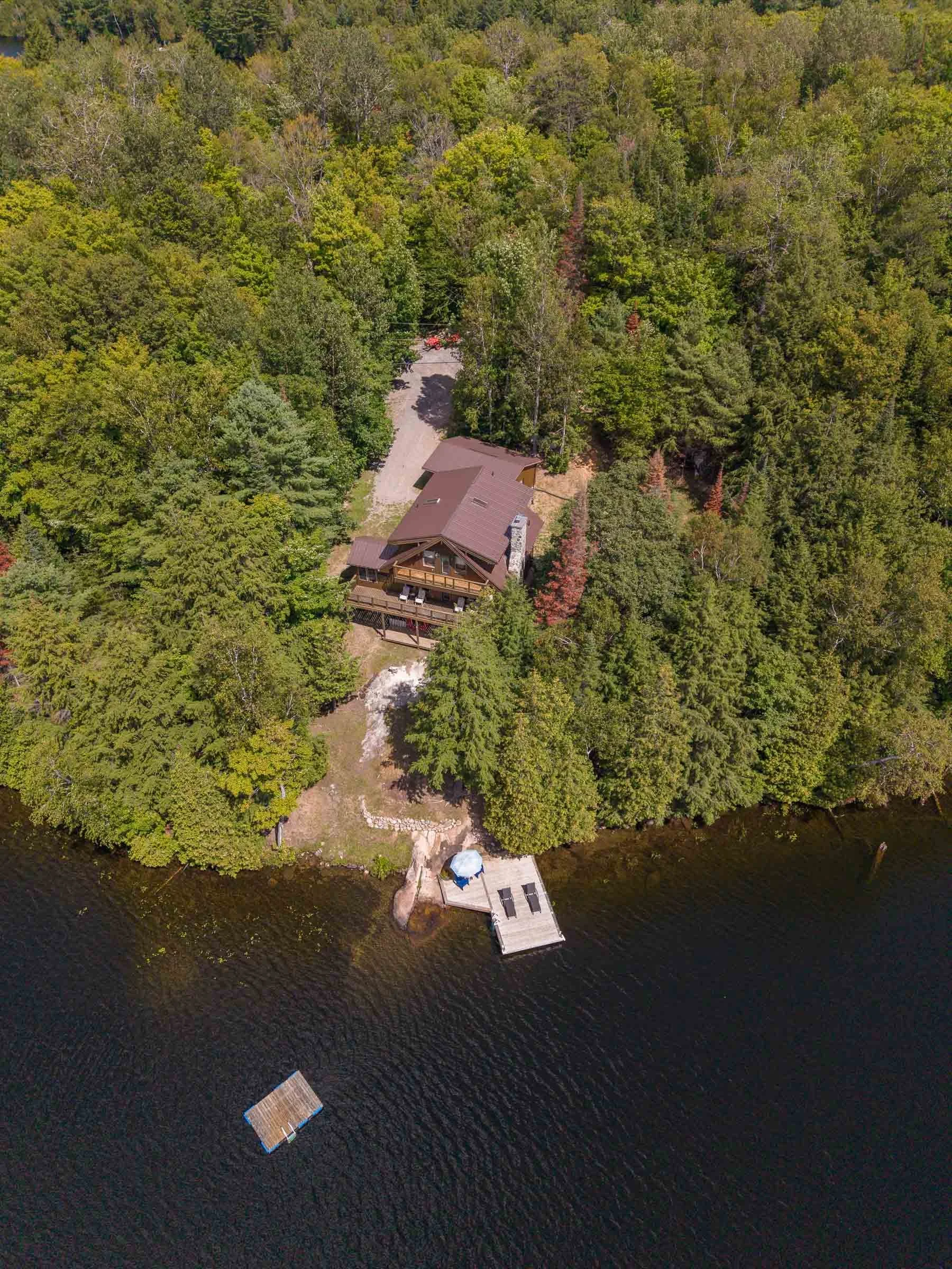 A house with a deck is situated among dense green trees near a body of water, with a small dock and two paddleboarders visible on the water.