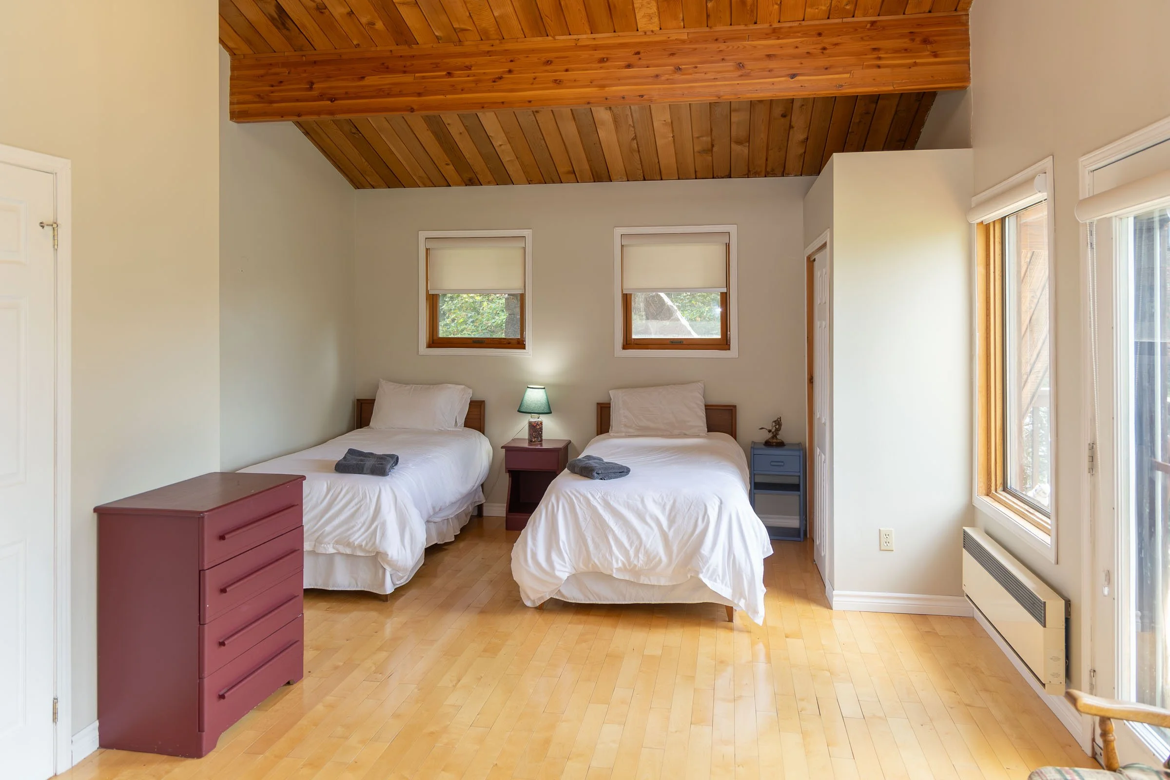 A bedroom with two single beds, each with white bedding and a gray towel, clothes, and small nightstands, one red and one blue, between the beds. There are three small windows with blinds behind the beds and a large window with blinds on the right wa