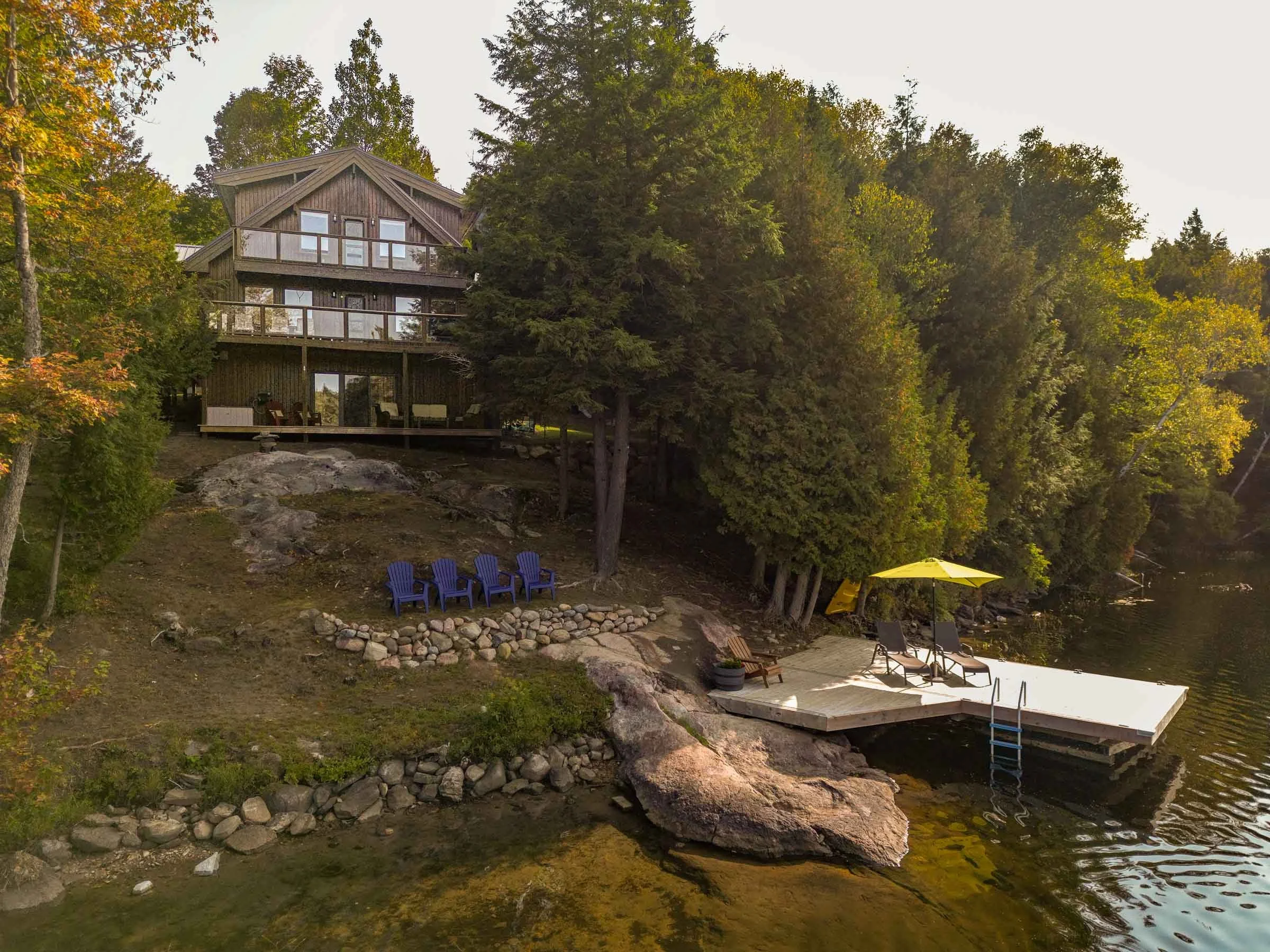 A wooden house with multiple decks situated among tall trees near a lake, with a dock, yellow umbrella, and outdoor furniture visible.