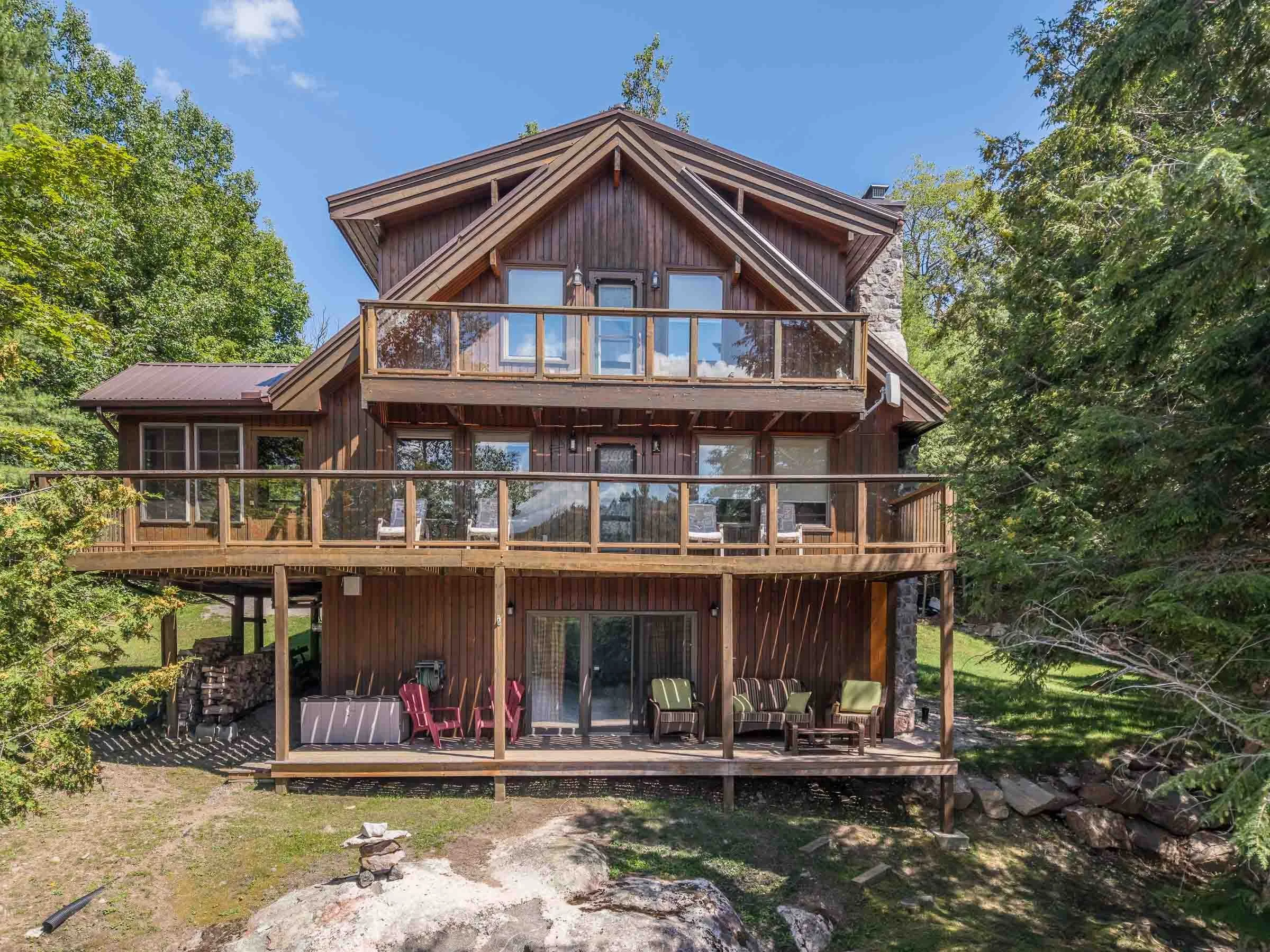 A multi-story wooden house with large windows and a balcony, surrounded by trees and set against a blue sky.