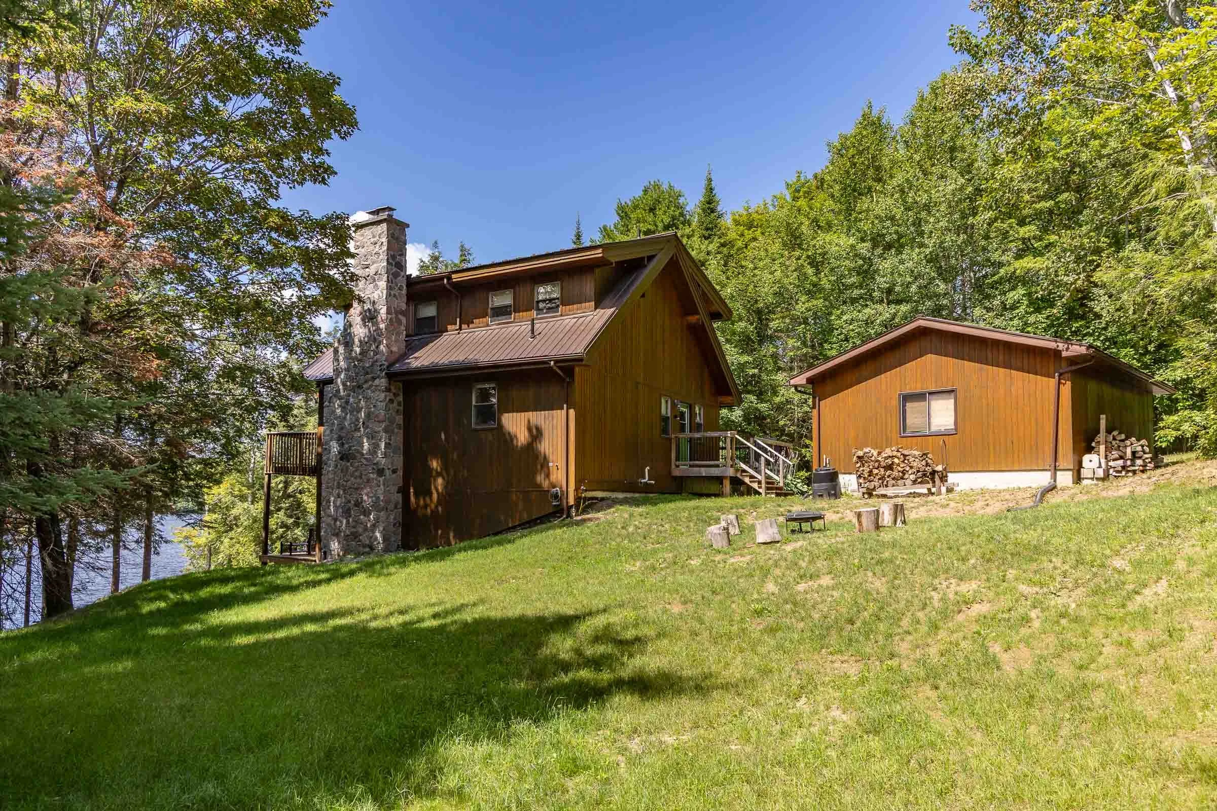 A wooden cabin with a stone chimney surrounded by green trees and grass, under a clear blue sky.