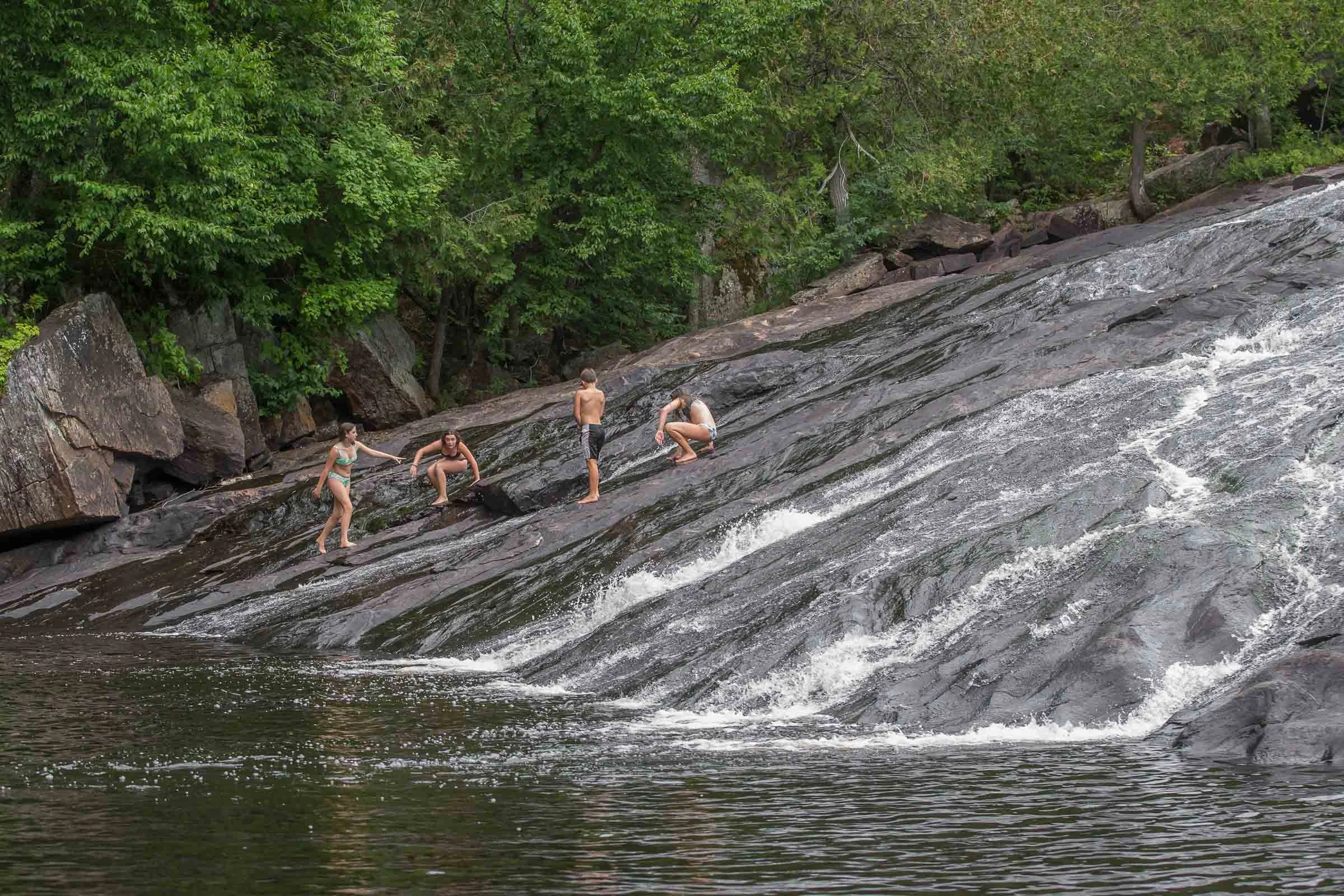 Four children playing and climbing on a rocky, sloped riverbank with flowing water, surrounded by green trees.
