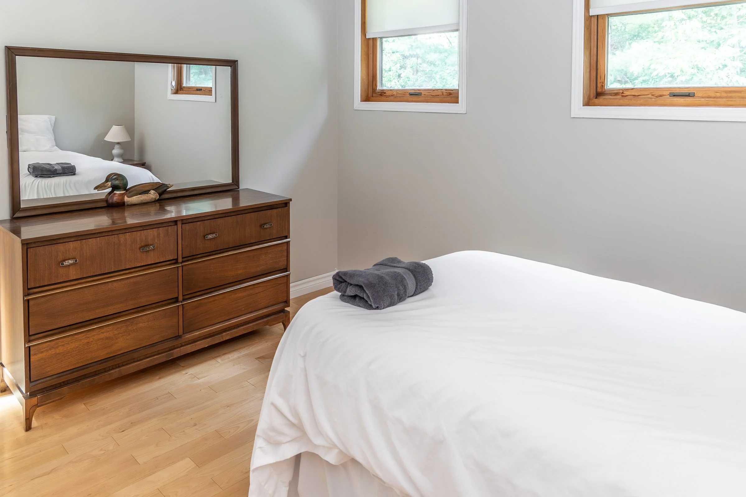 A bedroom with two beds, one with white linens and another with folded towels, wooden dresser with a mirror, and three windows with wooden frames showing greenery outside.