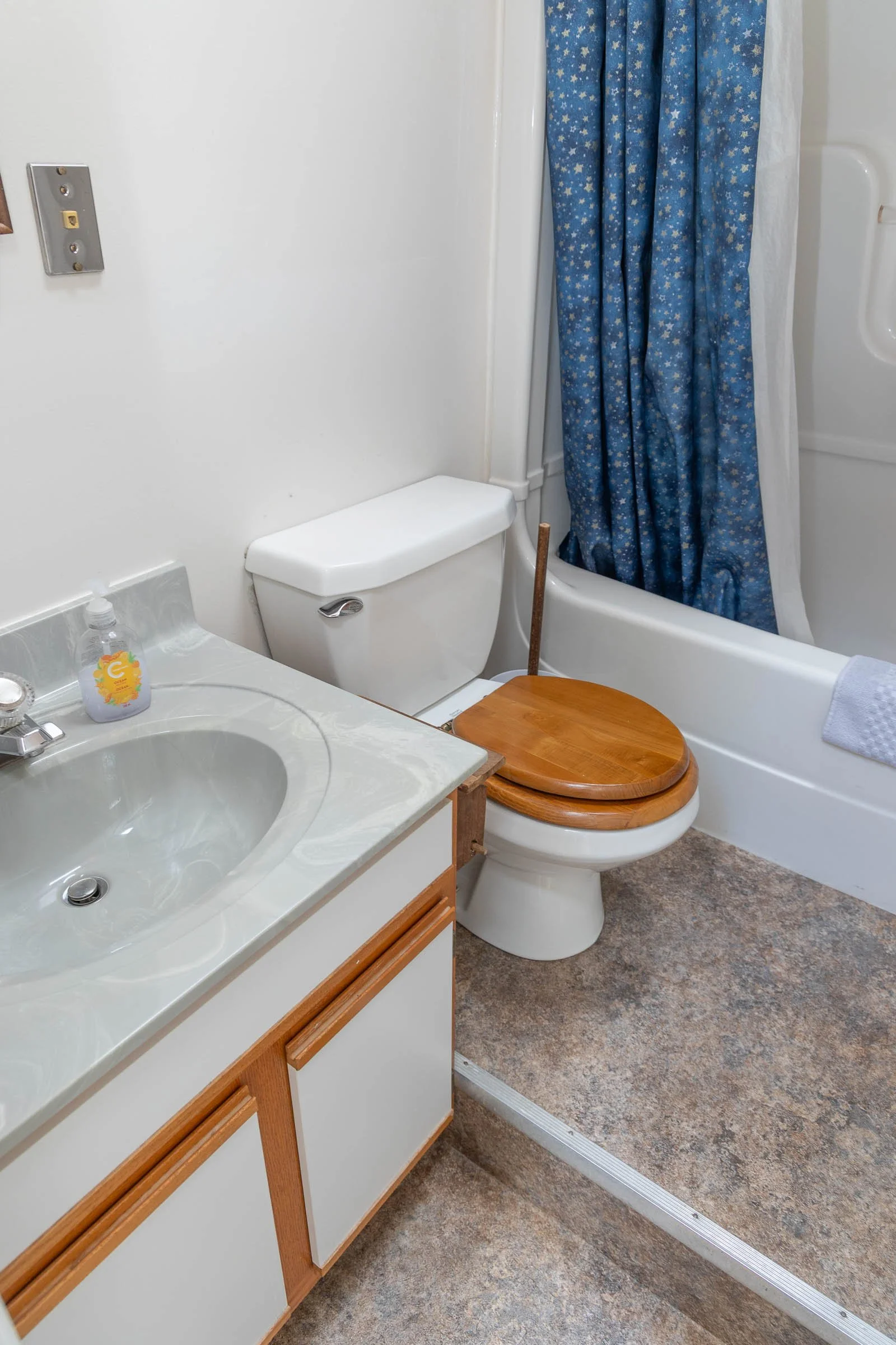 Bathroom with a toilet, a sink with a soap dispenser, a sink, a bathtub with a blue star-patterned shower curtain, a wooden toilet seat cover, and a metal shower track.