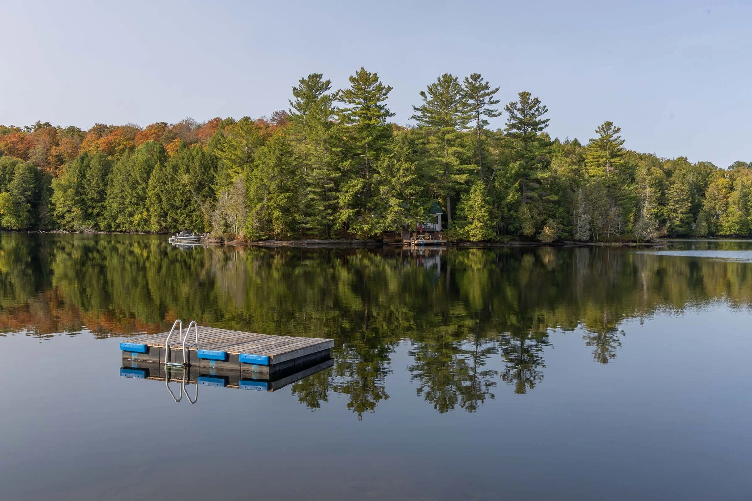 Calm lake with a wooden dock in the foreground and a house surrounded by green trees on the shoreline, reflected in the water during daytime.