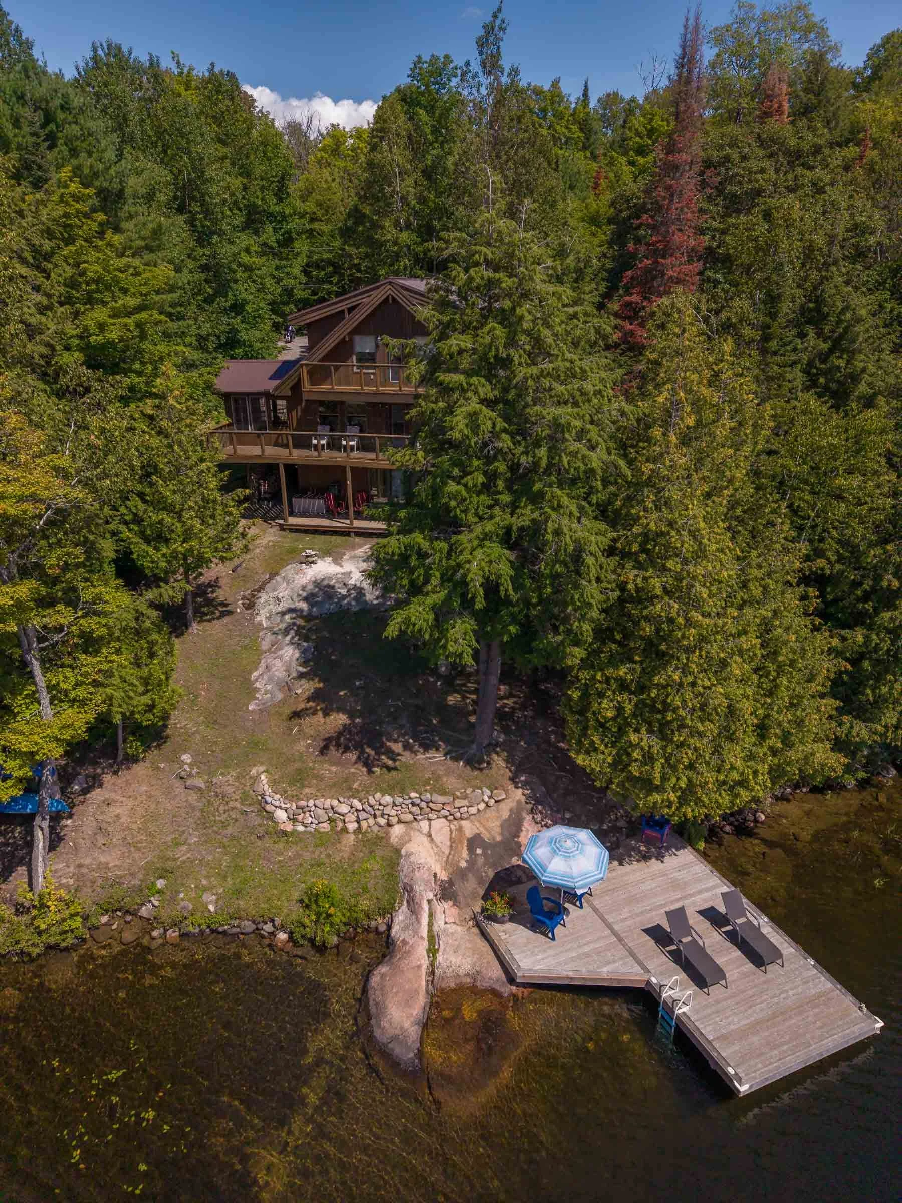 A house surrounded by trees with a deck extending over water, featuring outdoor furniture, an umbrella, and a small dock.