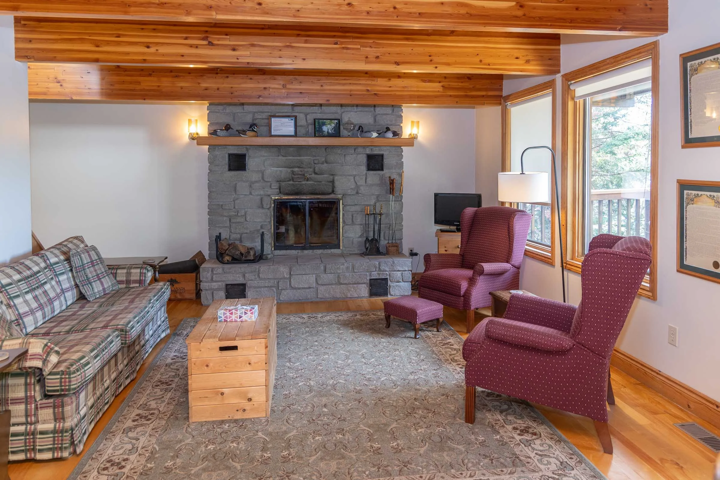 Living room with a stone fireplace, plaid sofa, red armchair, and large windows with wooden trim letting in natural light.