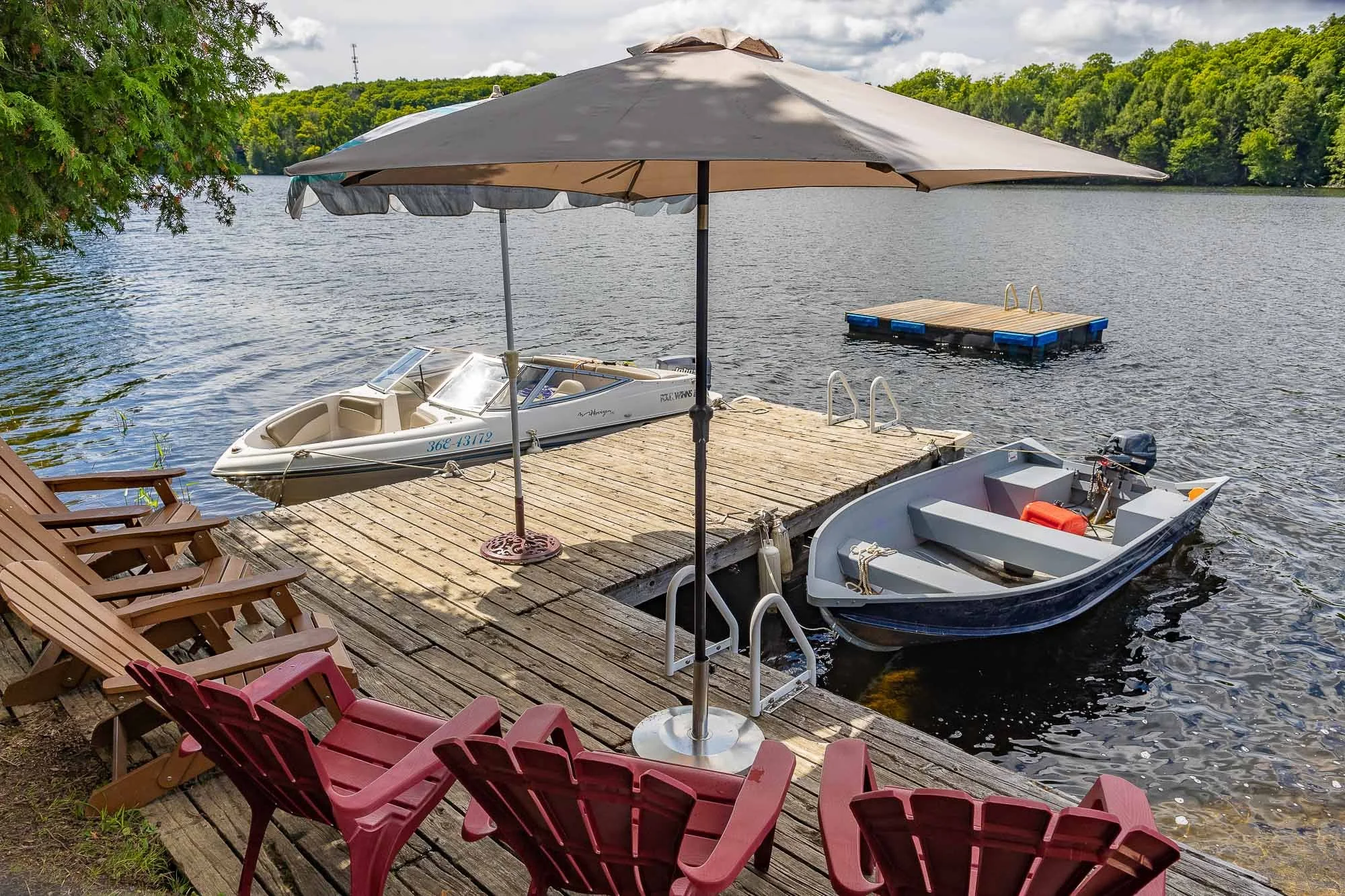 A lakeside dock with three boats tied to it, one with an umbrella providing shade, surrounded by lawn chairs in pink and red. The lake has a small floating dock and a larger dock in the background, with trees and hills on the opposite shoreline under