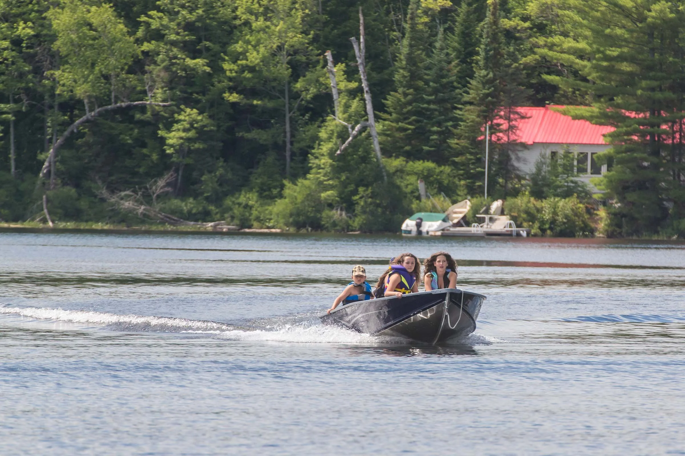 Four children wearing life jackets boating on a lake with green trees and a house with a red roof in the background.