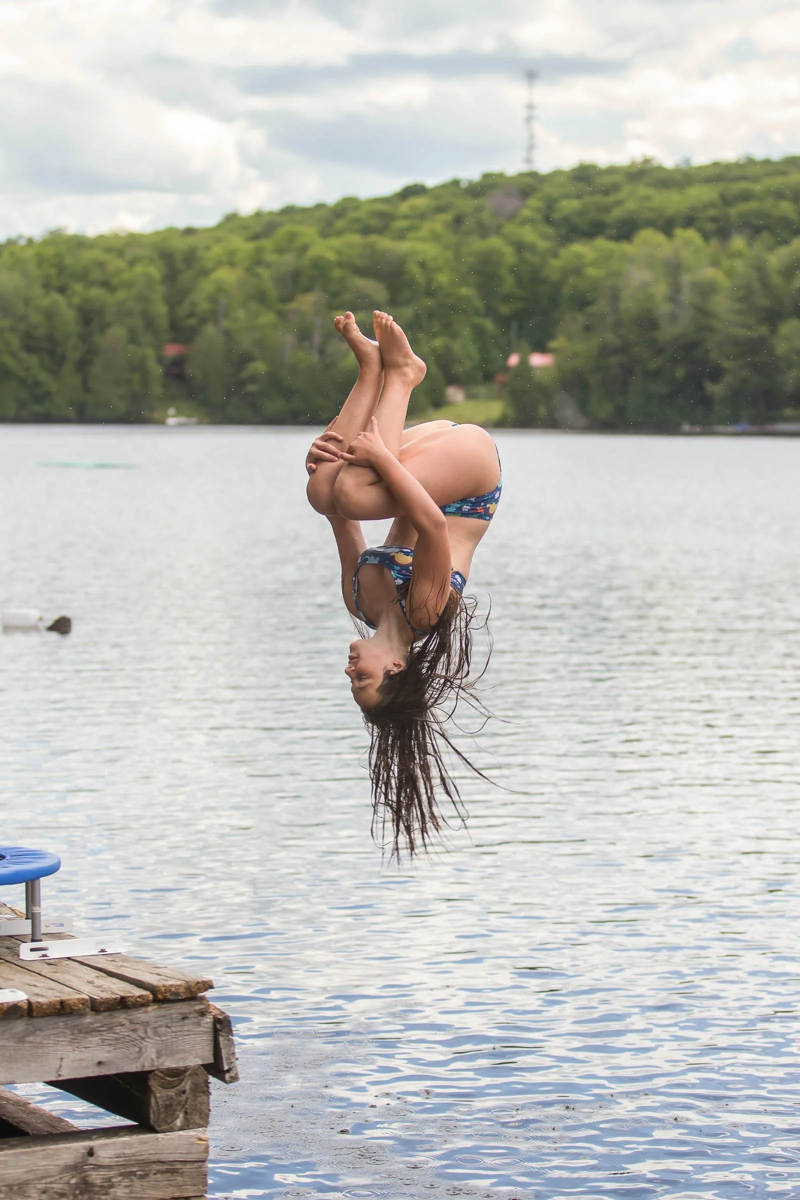A person in a swimsuit is flipping upside down into a lake from a wooden dock during daytime with water droplets mid-air, trees on the hillside, and a cloudy sky in the background.