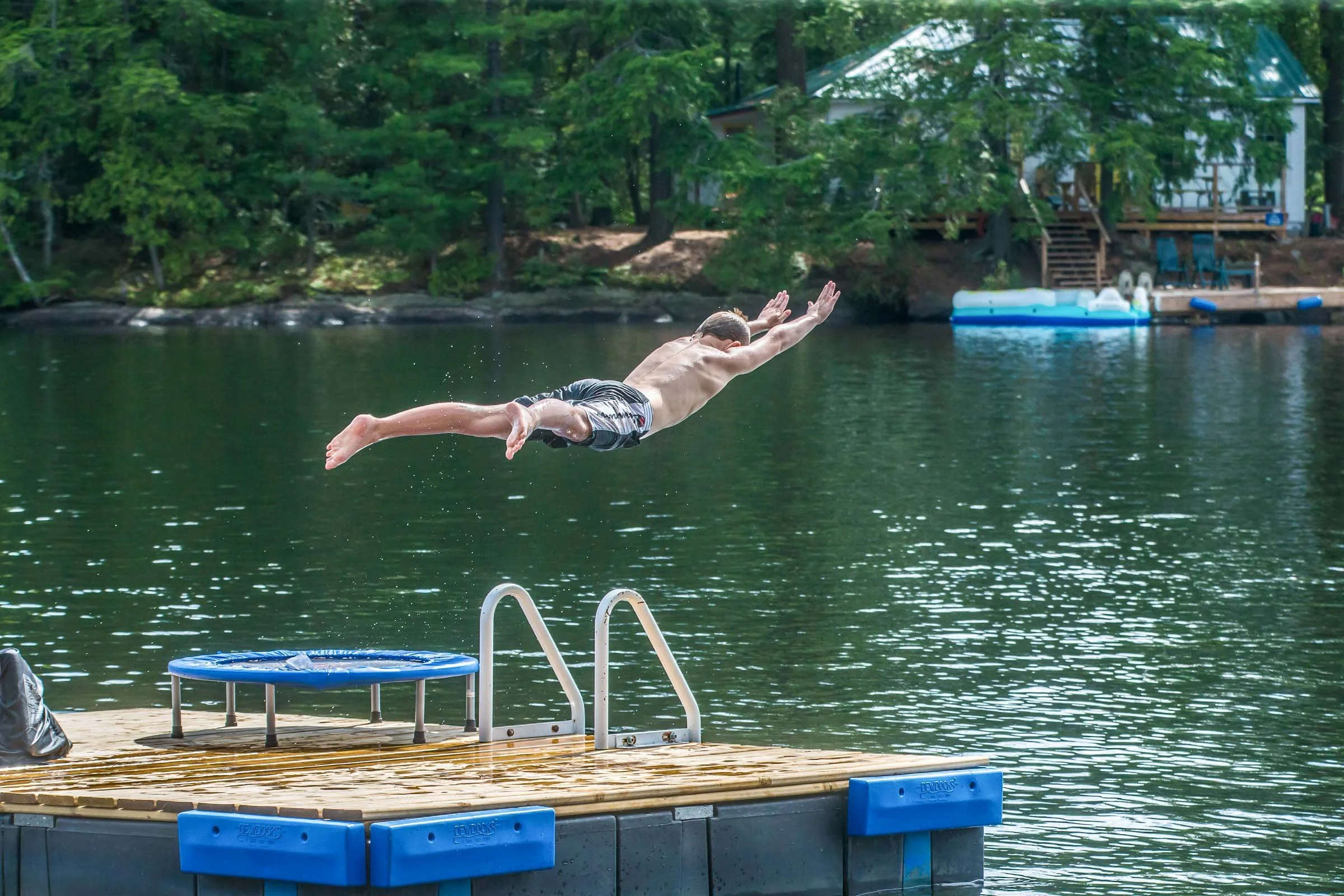 A man diving off a dock into a lake surrounded by trees with a house and an inflatable boat in the background.