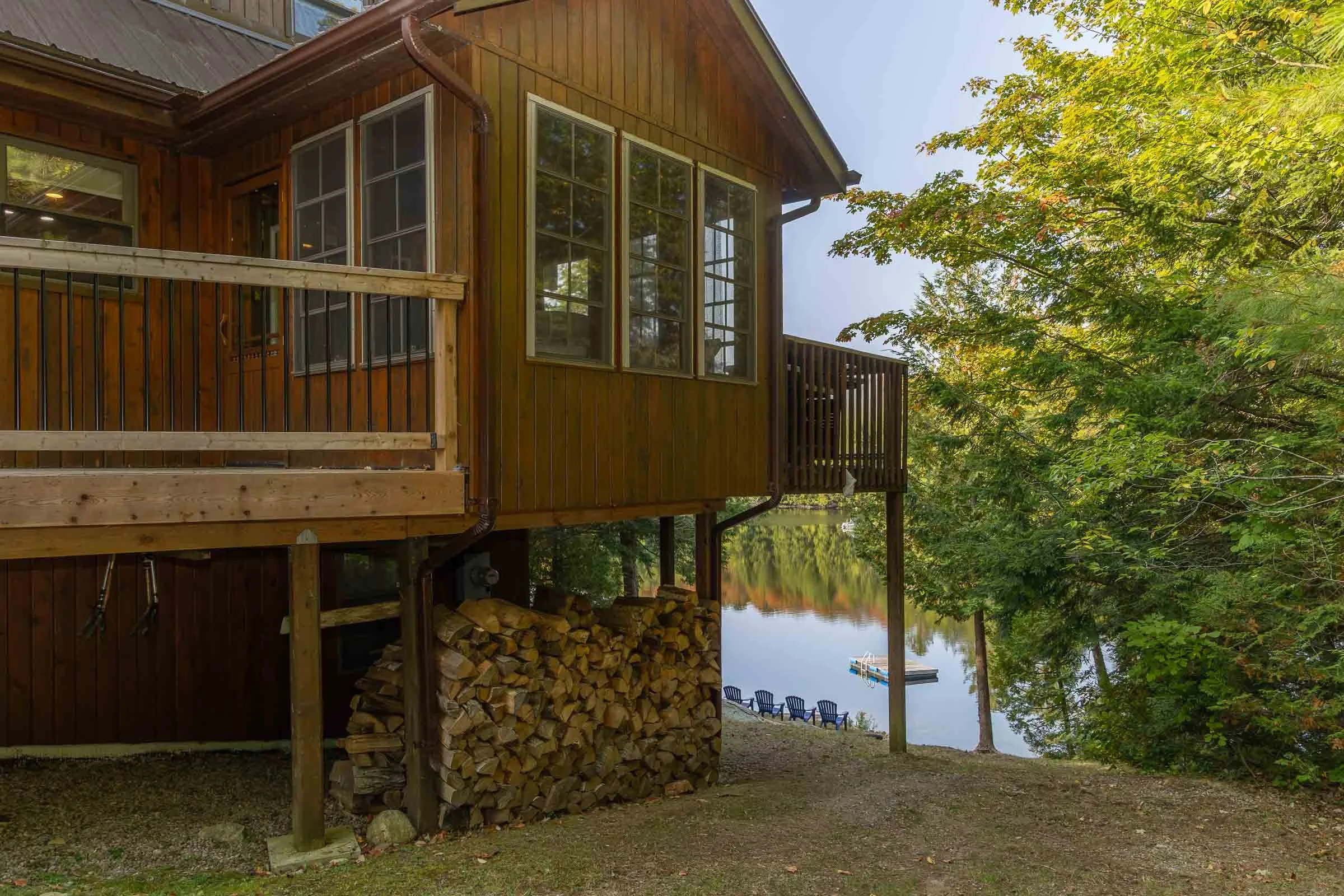 A wooden cabin near a body of water with a small deck, stacked firewood underneath, and trees with green leaves in the surroundings.