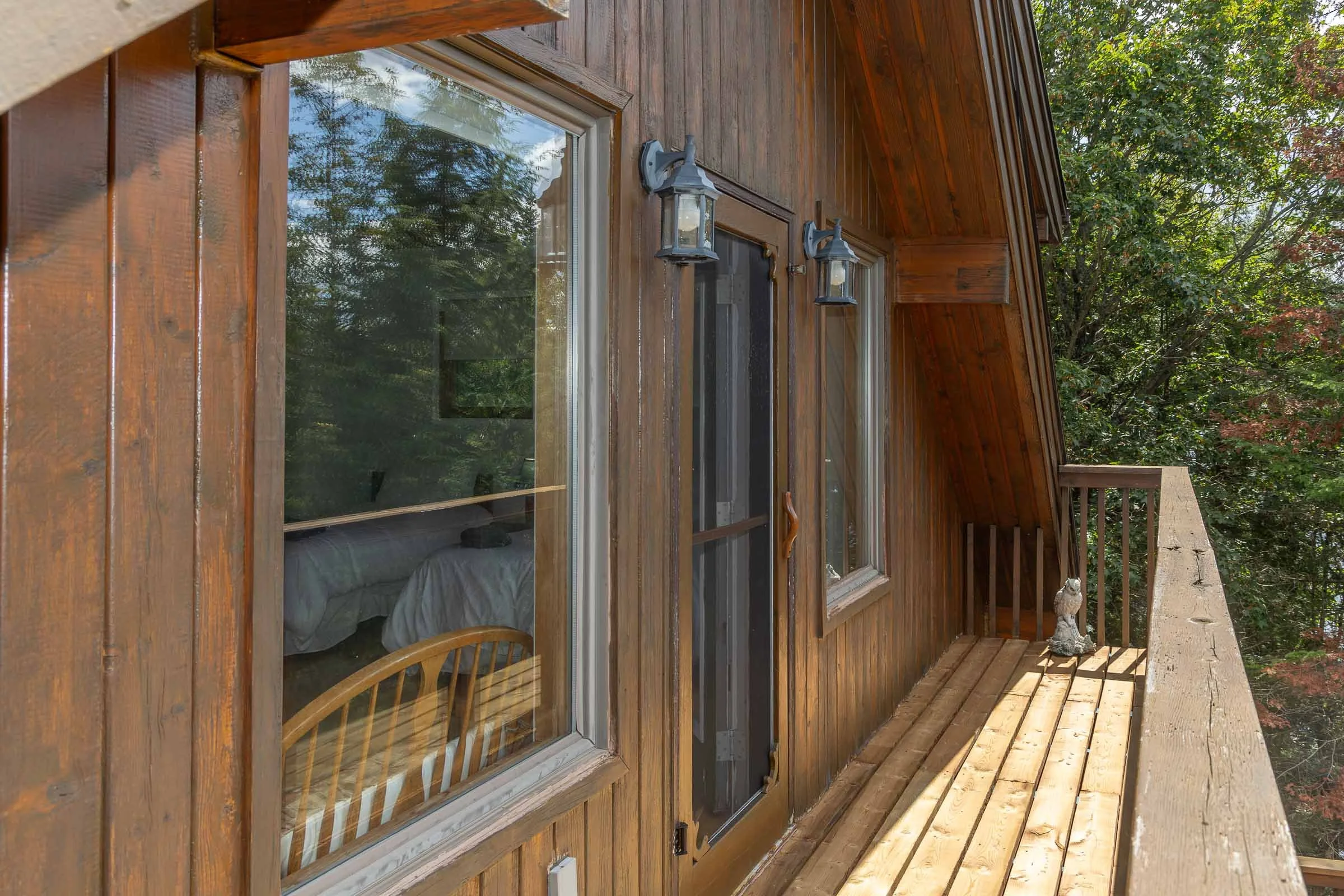 A wooden house exterior with a large window showing a bedroom inside, two outdoor lanterns mounted on the wall, and a small deck area surrounded by green trees.