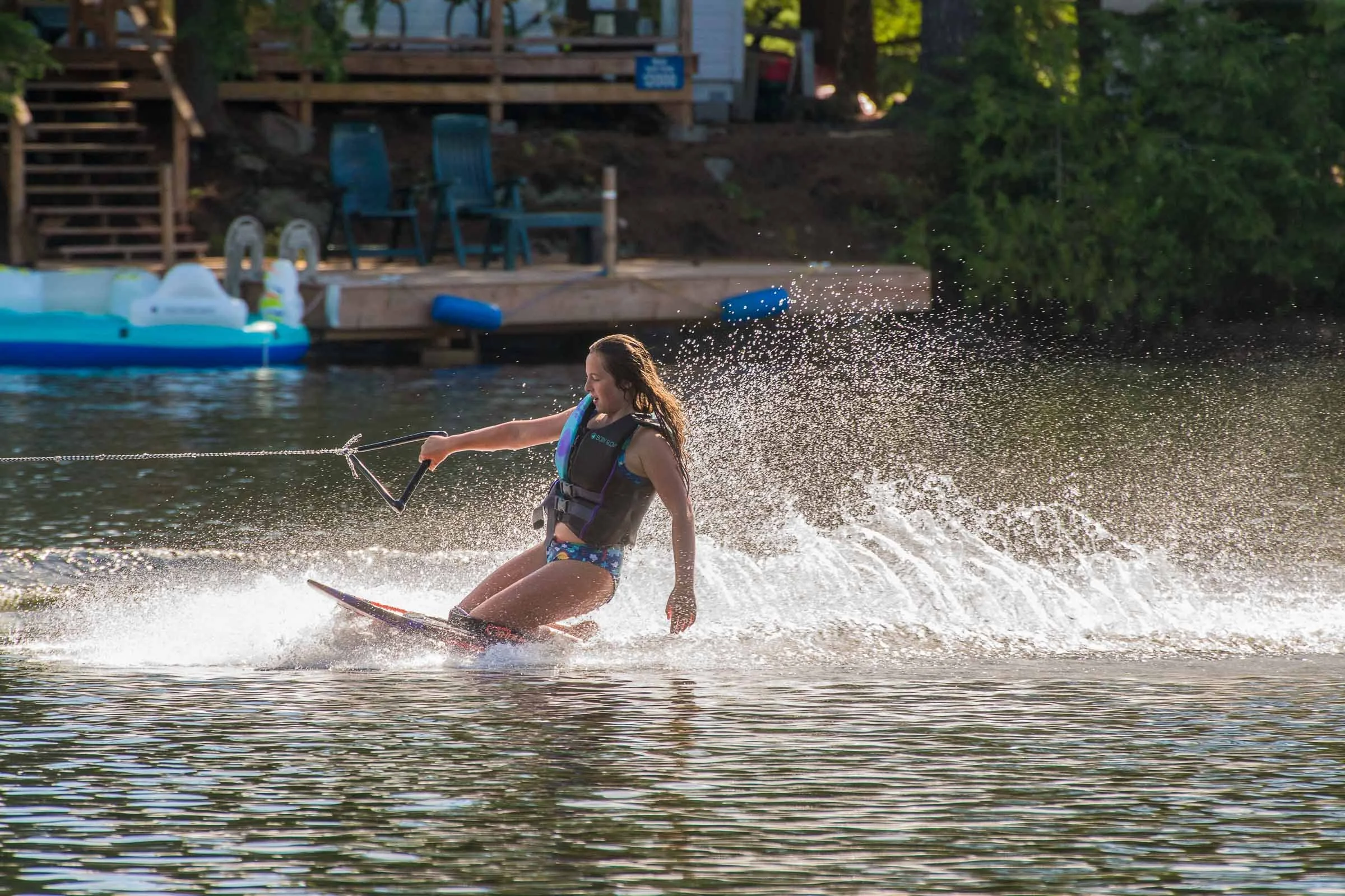 A young woman wearing a life jacket, a swimsuit, and sunglasses is wakeboarding on a lake, creating a spray of water behind her as she rides the water.