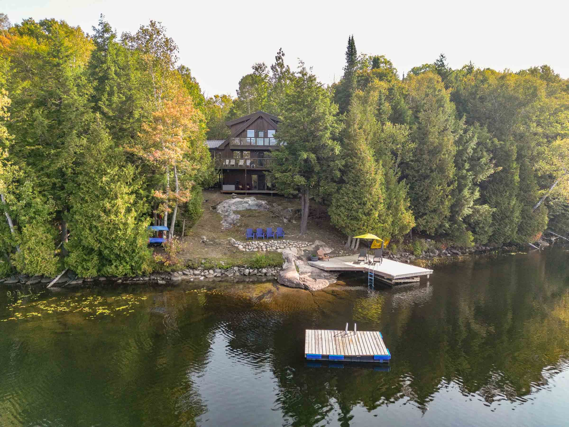 A wooden house surrounded by lush green trees on a hillside, overlooking a lake with a floating dock and several Adirondack chairs, with a small yellow canopy on the right side.
