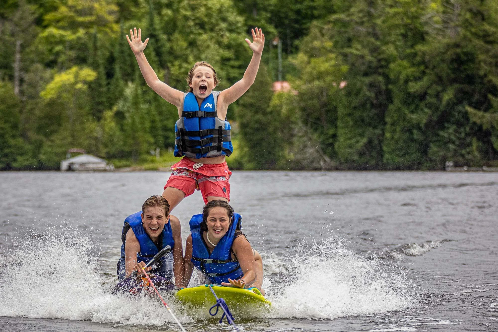 Four children wearing life jackets are on a yellow rubber raft and appear to be enjoying a boat ride on a lake or river during daytime, with a forested area in the background.