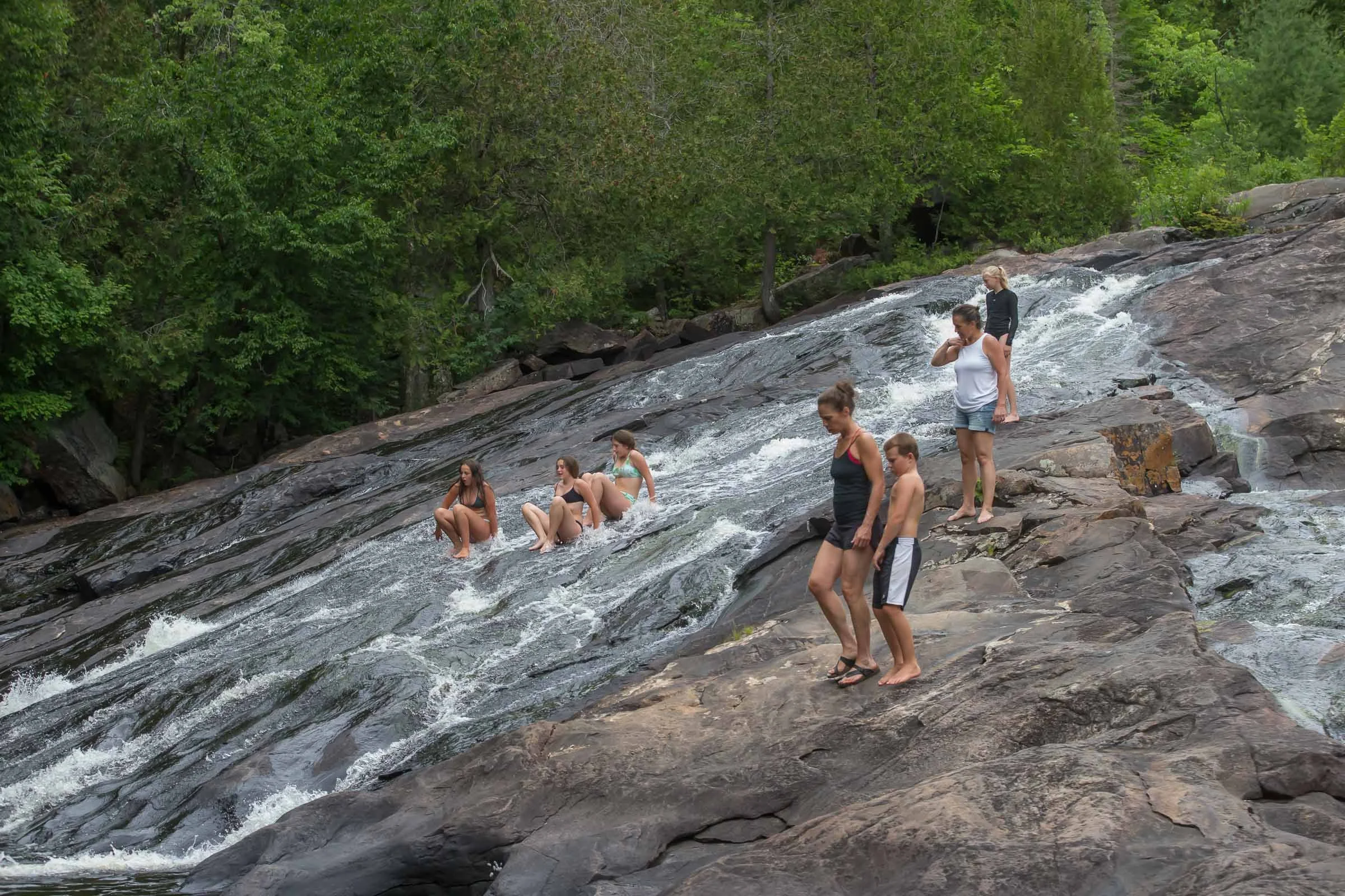 People cooling off and sitting on rocks in a shallow mountain stream surrounded by green trees.