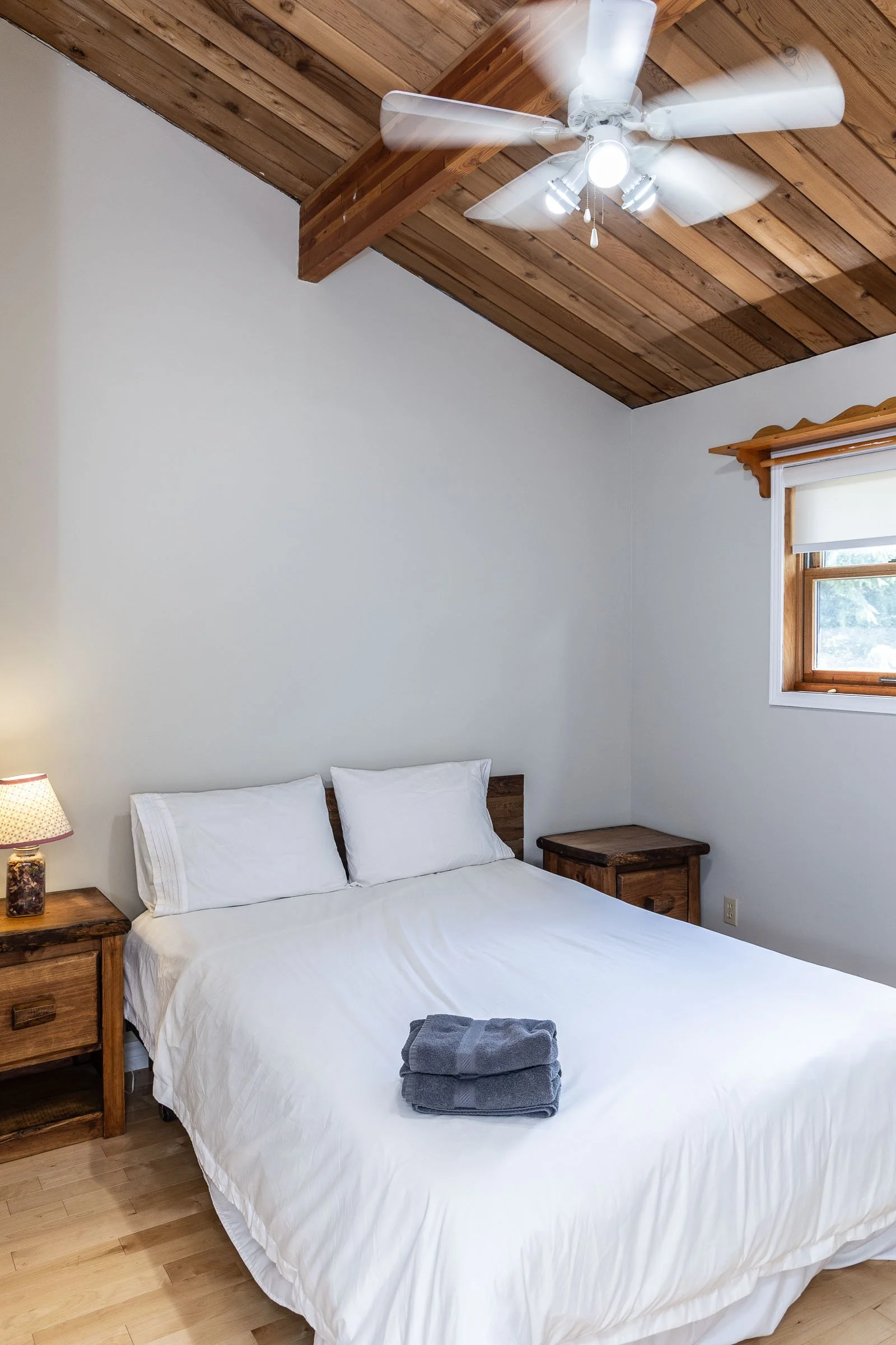 A bedroom with a white bed, two pillows, and towels on it. Two wooden nightstands on each side of the bed. A small lamp on the left nightstand. The ceiling has wood paneling and a ceiling fan with lights. A window with a wooden frame and a partially 