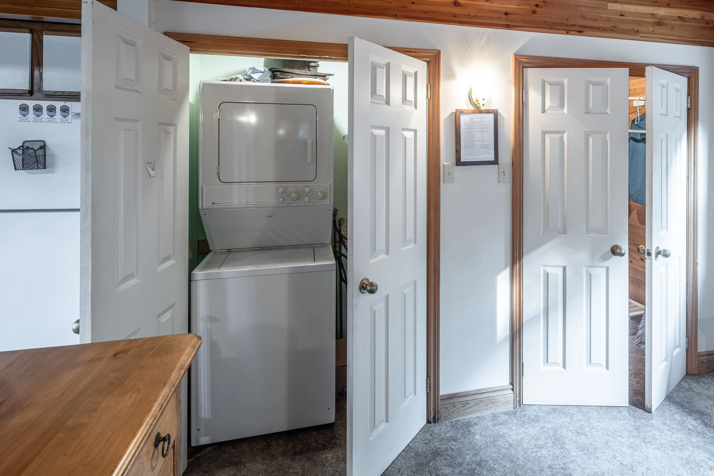 A small laundry room with a white stacked washer and dryer unit, partially hidden behind a white door. The room features wood trim around the doors and ceiling, with a wooden table in the foreground and a beige carpeted floor.