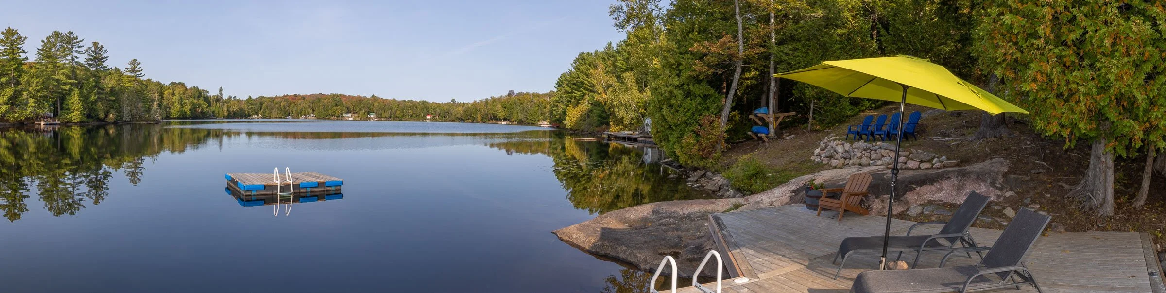 Calm lake with reflections of trees and a clear sky, surrounded by green foliage. A floating dock with a ladder is on the water. The shoreline has a wooden deck with lounge chairs, a yellow umbrella, and Adirondack chairs under trees.