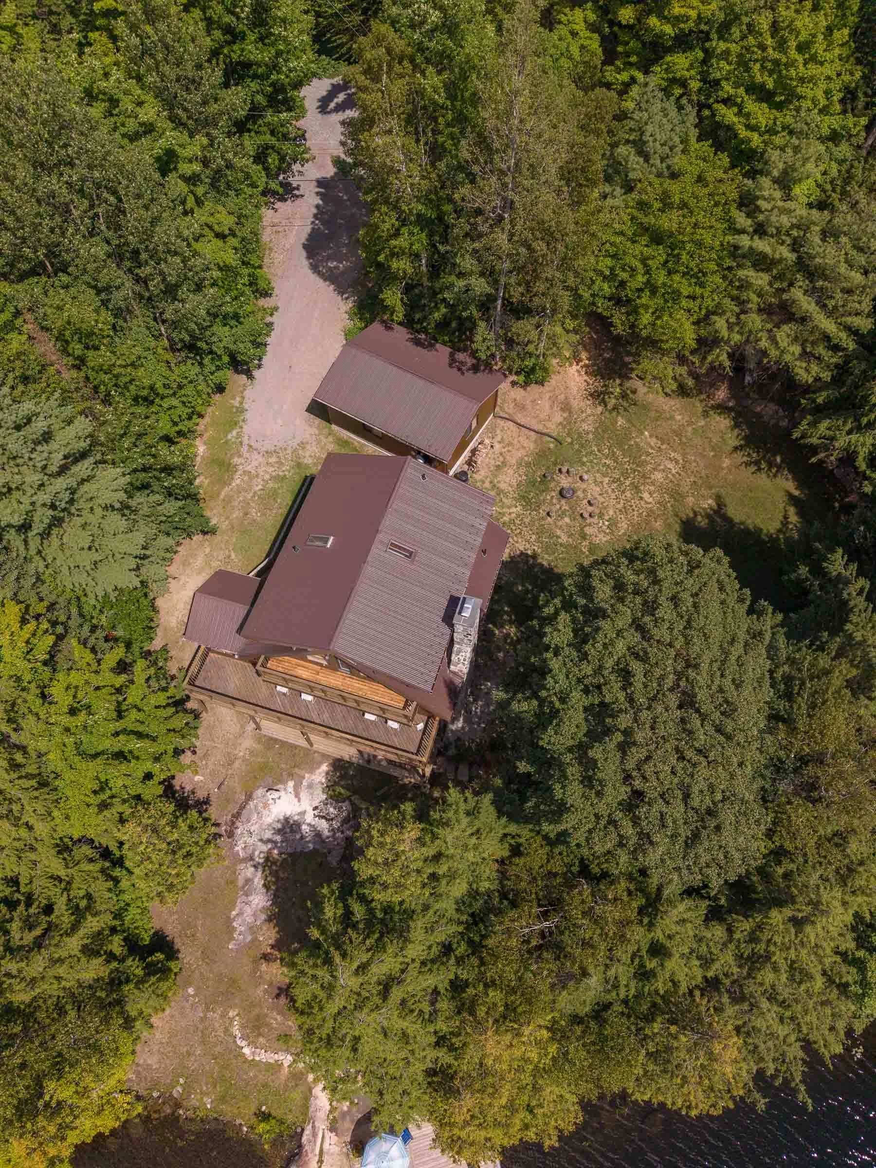 An aerial view of a house surrounded by lush green trees, with a gravel driveway and a small creek running nearby.