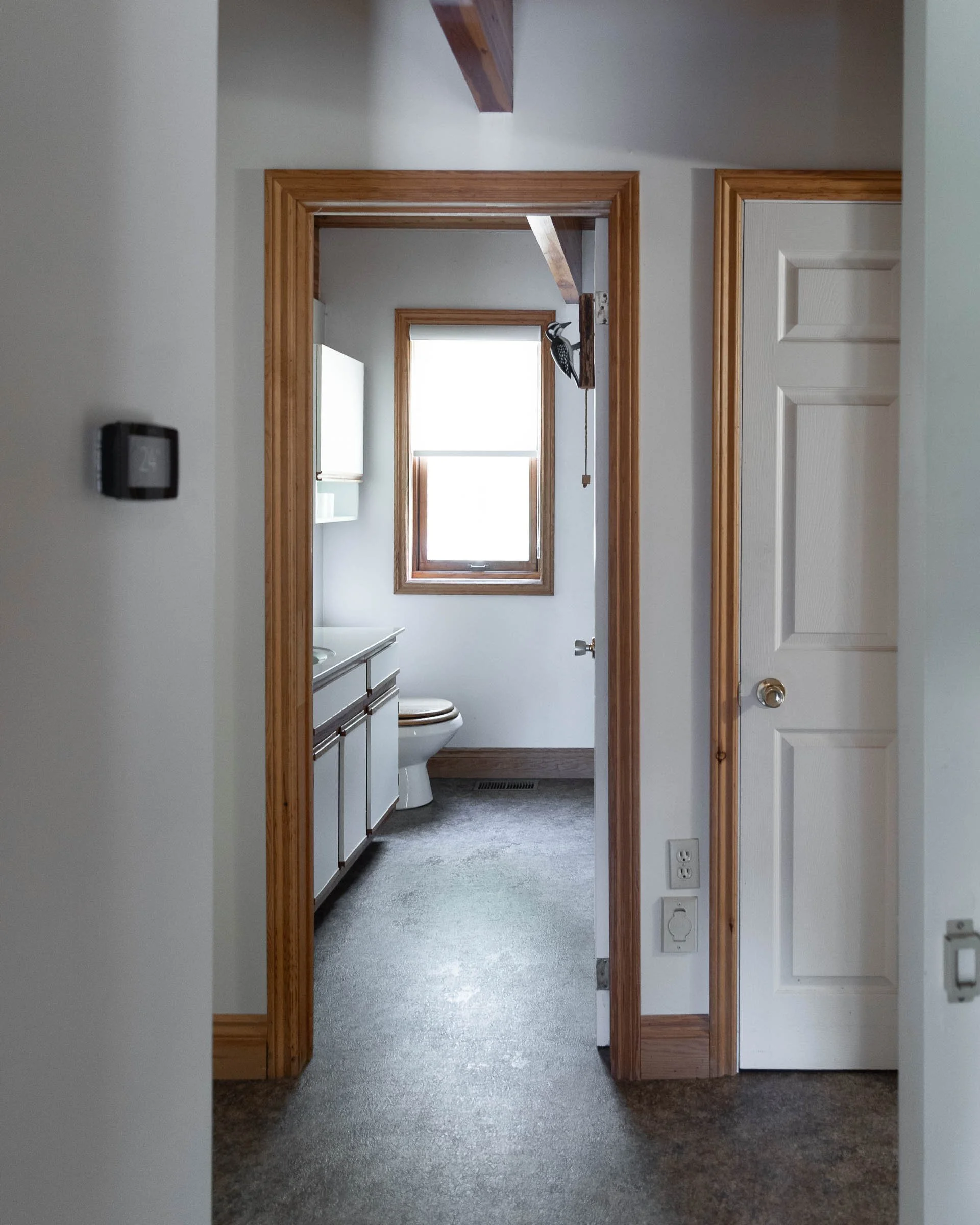 A hallway view leading into a bathroom with a toilet and window, surrounded by wooden door and window frames, carpeted hallway floor, and wall outlets.