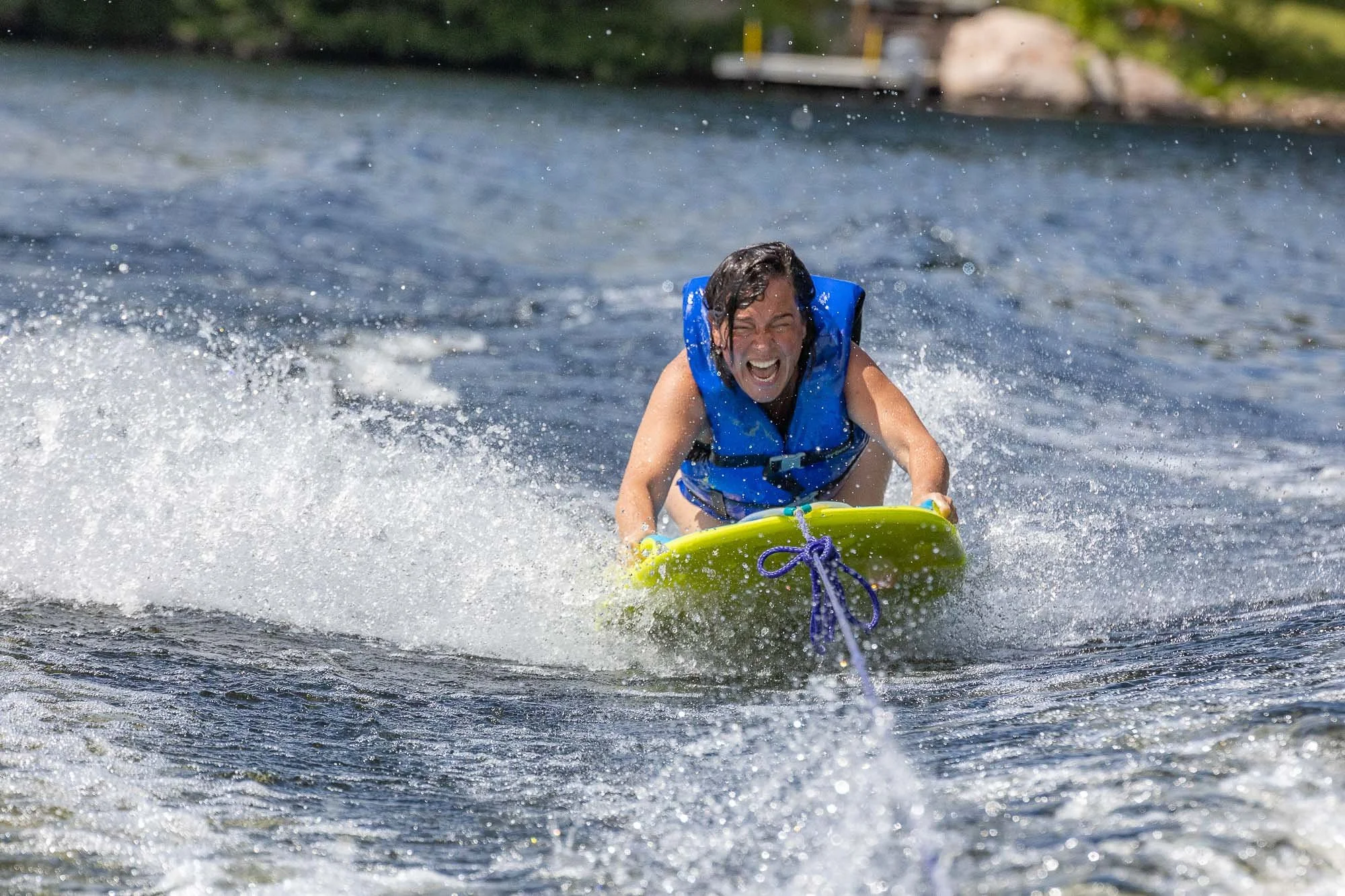 Person wearing a blue life jacket riding a wakeboard on a lake, smiling and splashing water.
