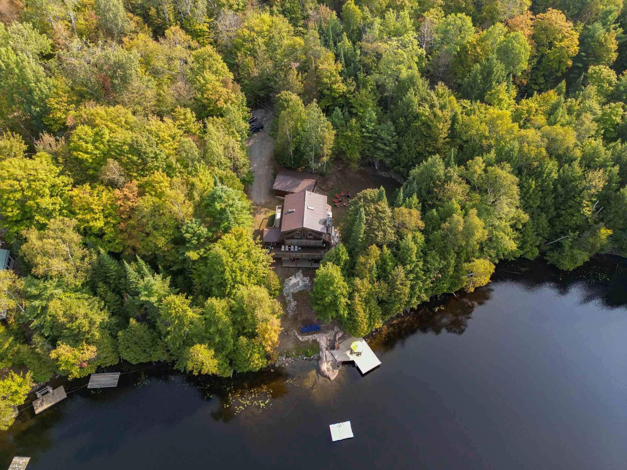 An aerial view of a house surrounded by dense green trees with a shoreline and water body nearby, with several docks and boats visible along the water's edge.