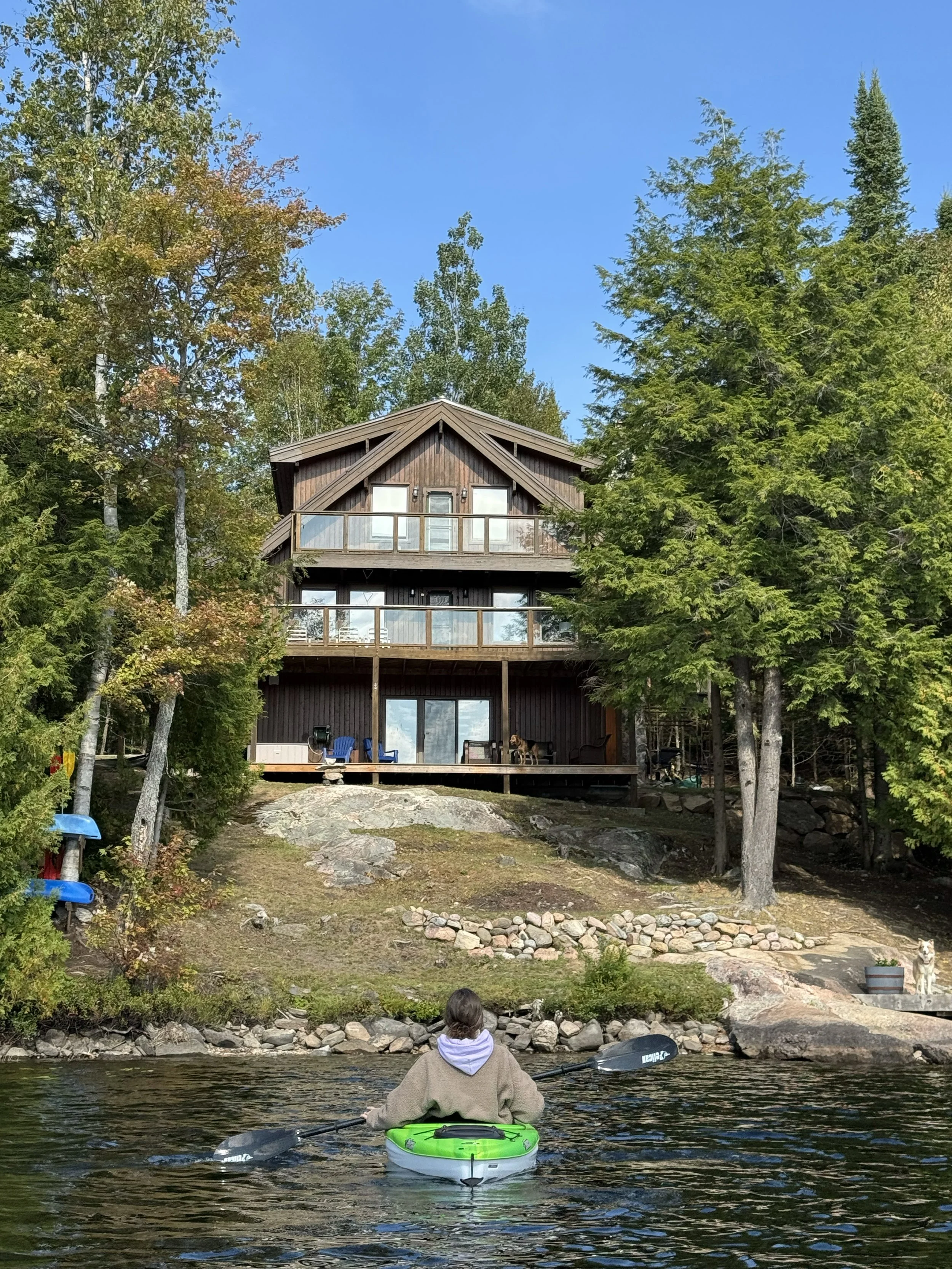Person sitting in a kayak on a river, facing a house on a hill surrounded by trees.