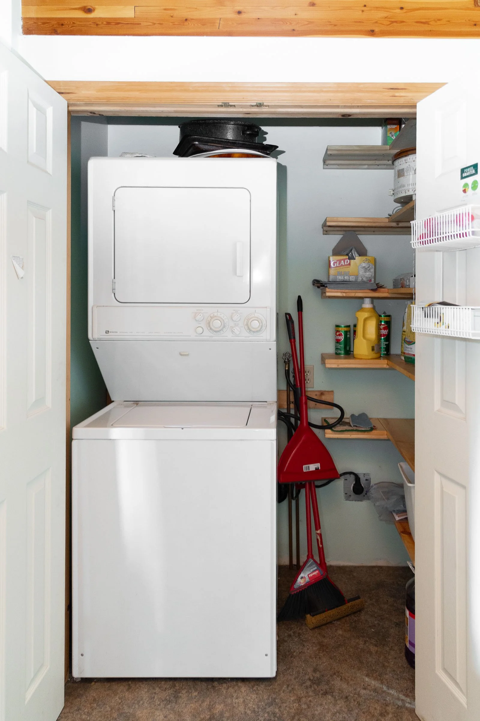 View of a small laundry closet with a white stacked washer and dryer, wooden shelves holding cleaning supplies, a broom, dustpan, and various stored items. The closet is framed by partially open white double doors.