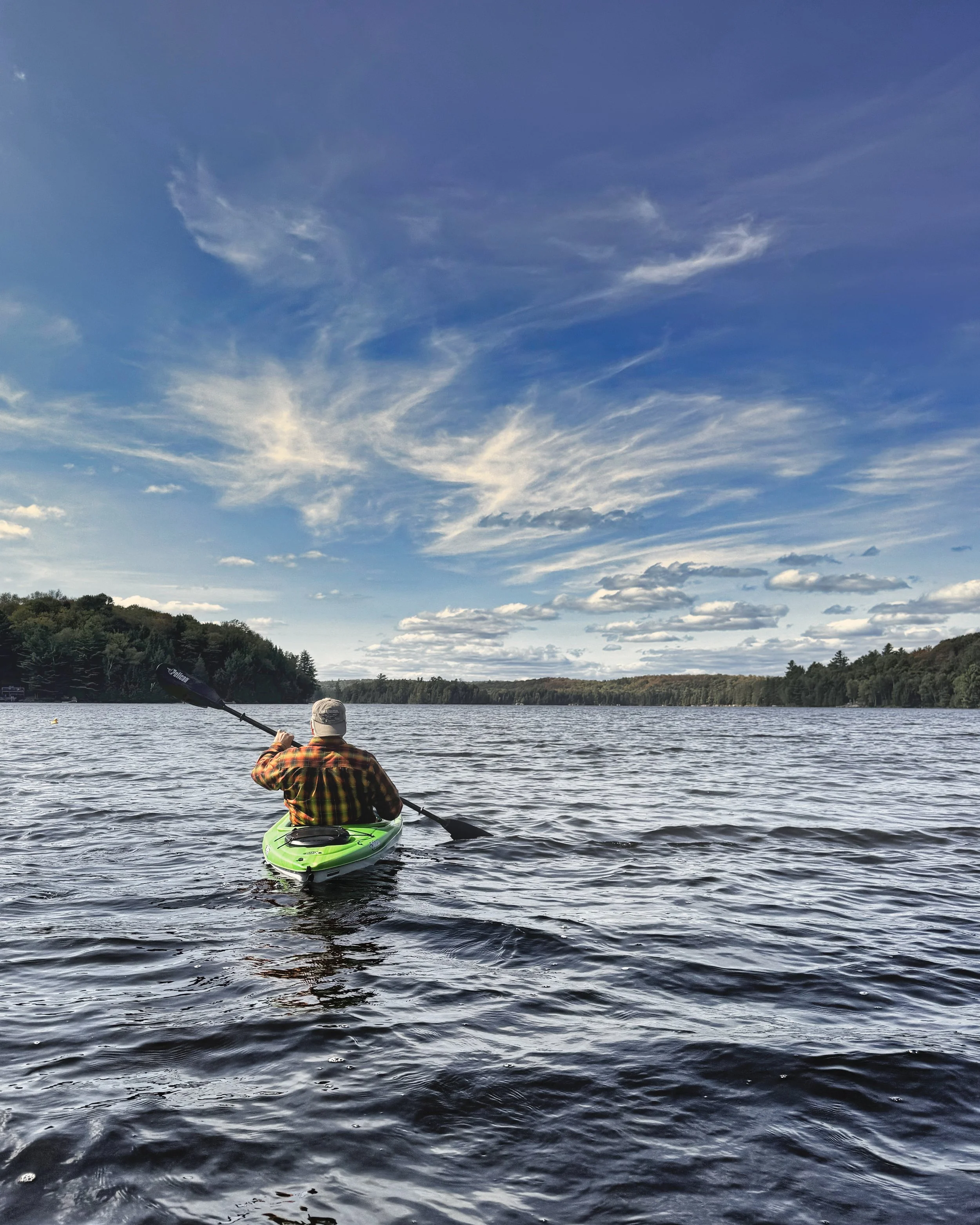 Person kayaking on a body of water under a partly cloudy sky, with trees along the shoreline in the background.