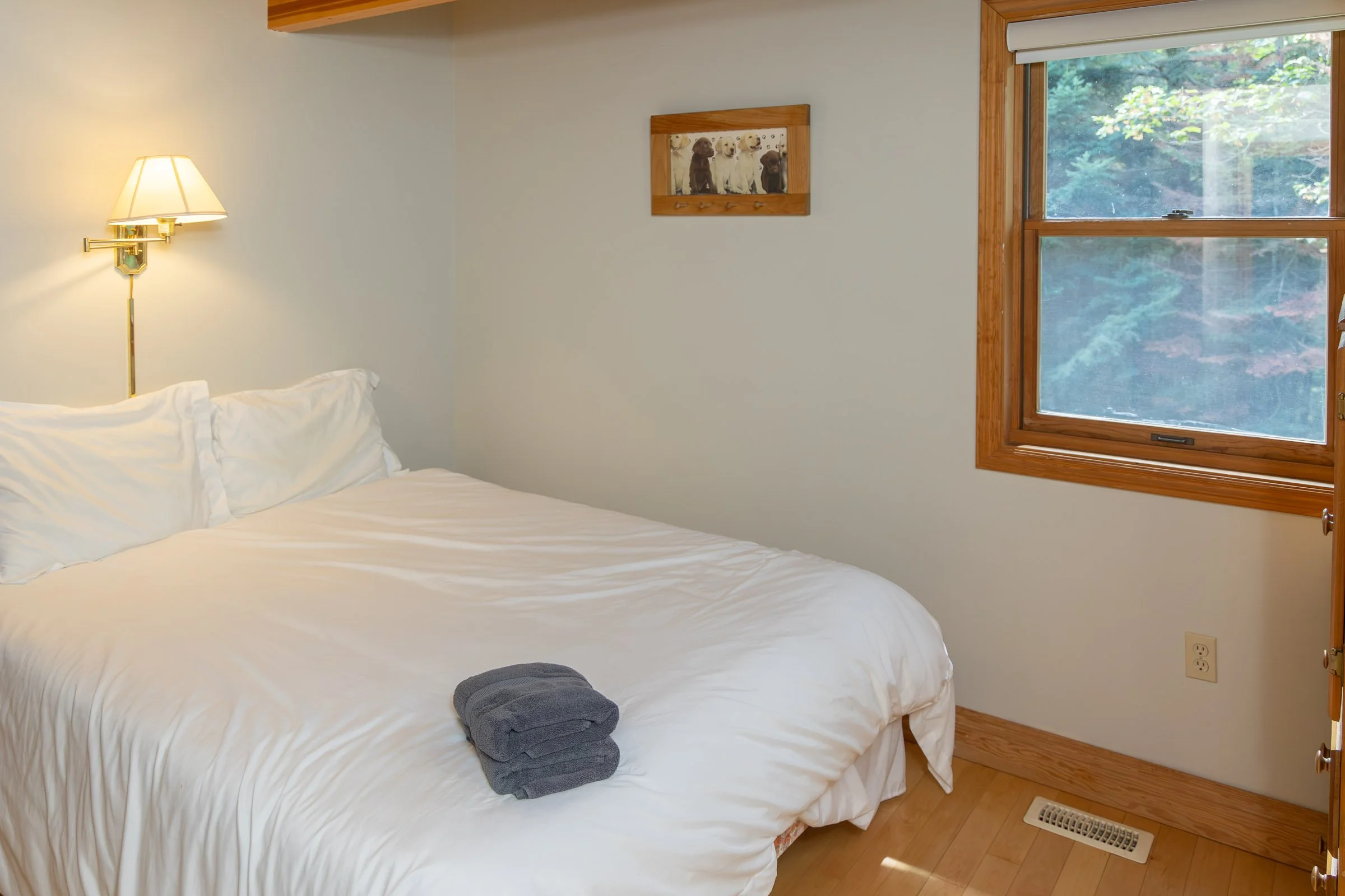 A neatly made bed with white sheets and pillows, a small rolled-up gray towel at the foot, situated in a room with cream-colored walls, a wood-framed window showing green foliage outside, and a wall-mounted light fixtures with cream shade above the h