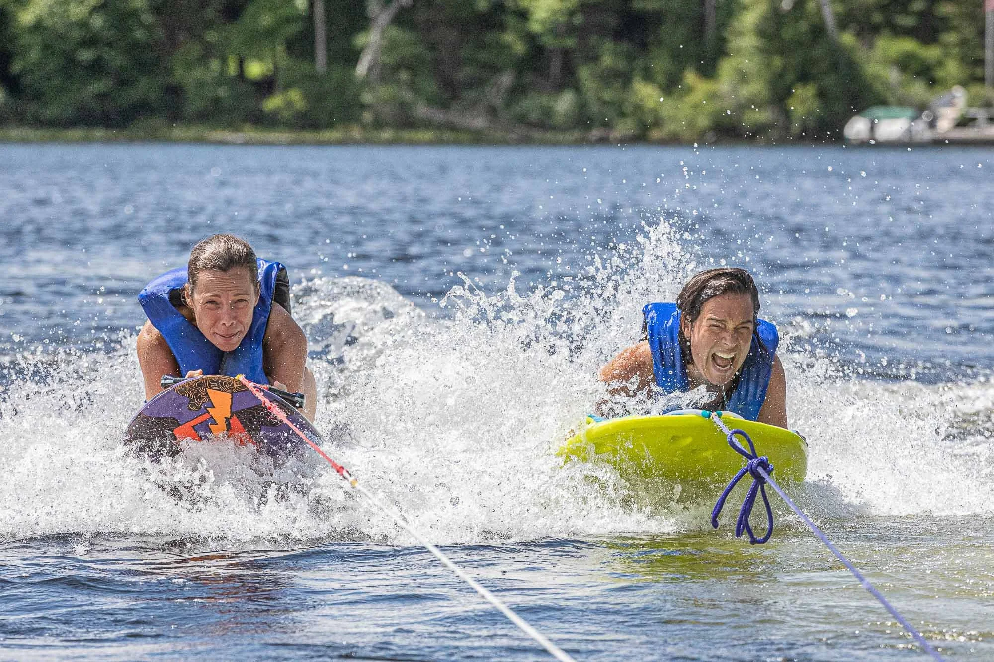Two women in life jackets are water skiing on a lake, holding onto a tow rope with trees and a boat in the background.