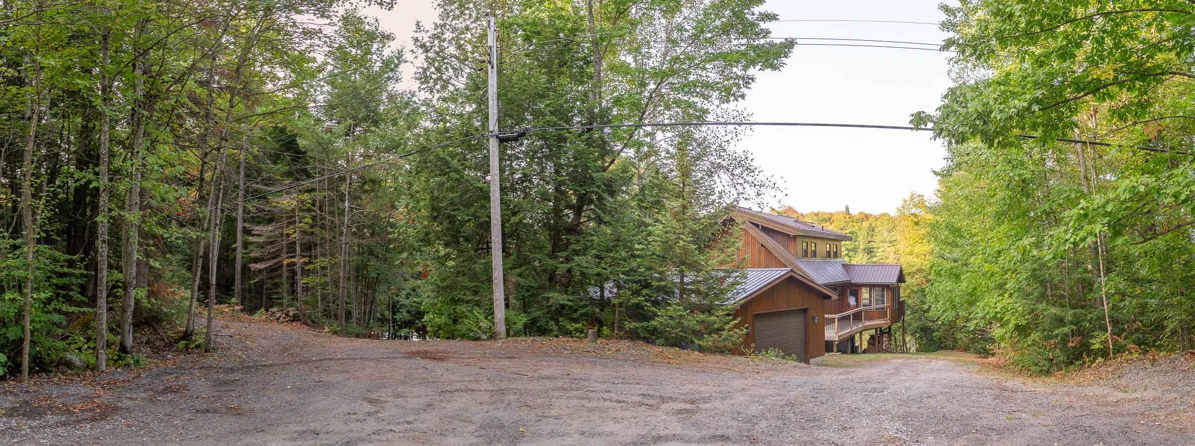A house nestled among trees in a wooded area with a gravel driveway and electrical lines overhead.