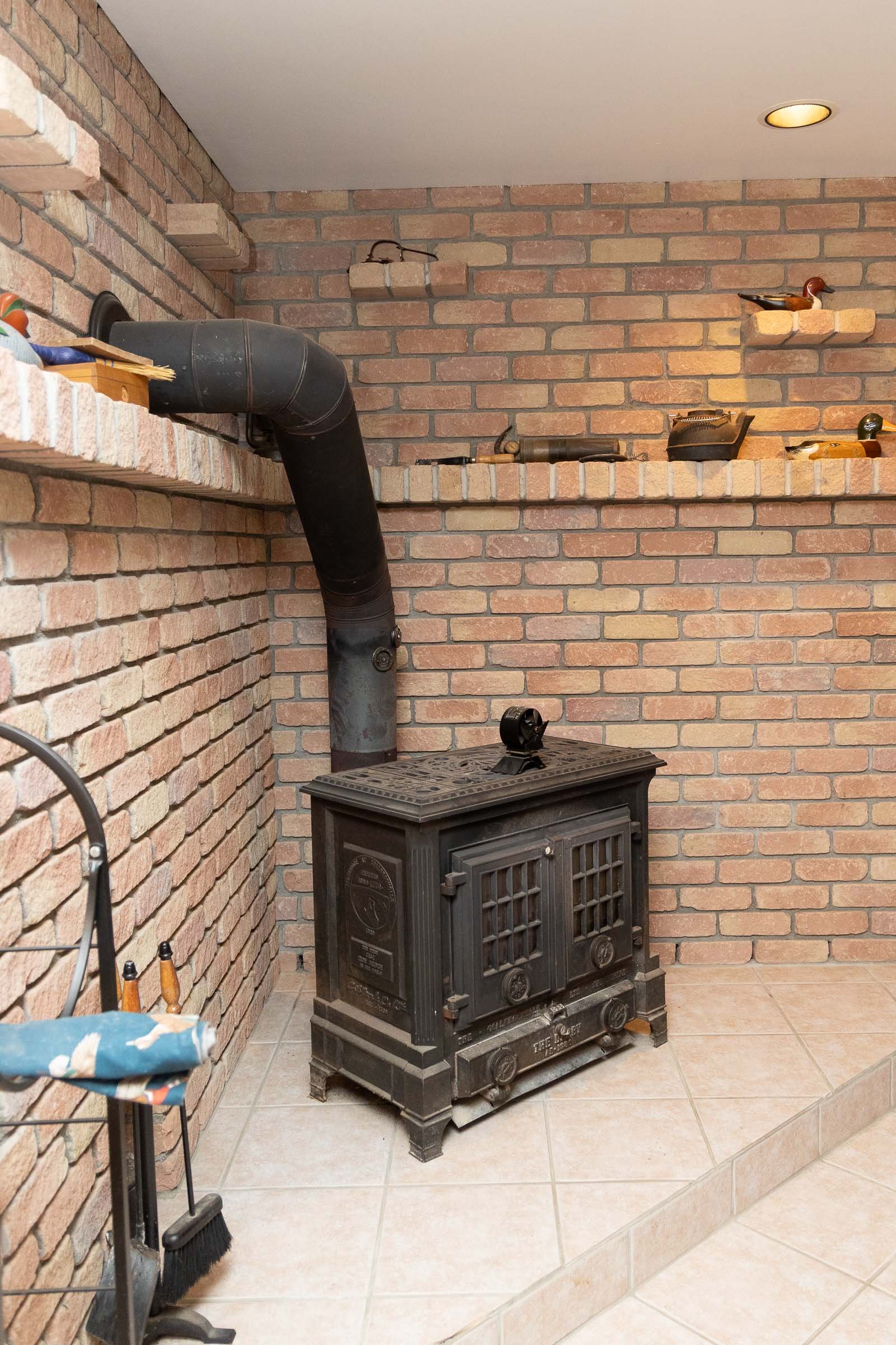A vintage black cast-iron stove placed on a tiled floor against a brick wall in a room with a sloped ceiling. There are decorative ducks on a shelf, various tools, and a chimney pipe connected to the stove.