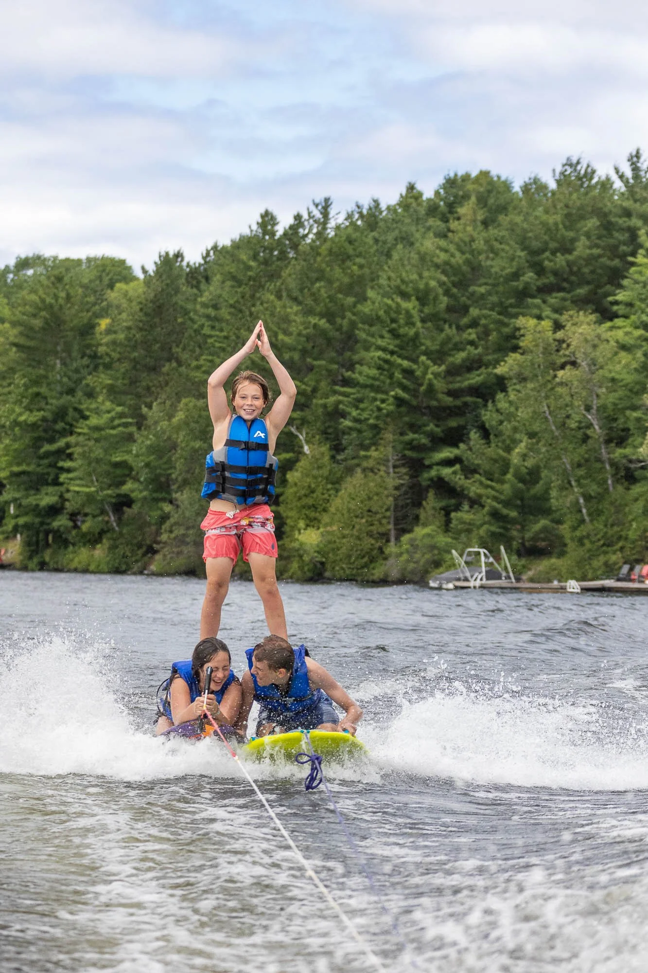 Group of children wearing life jackets on a float, with one girl standing on top, in a lake with a forested shoreline and a boat in the background.