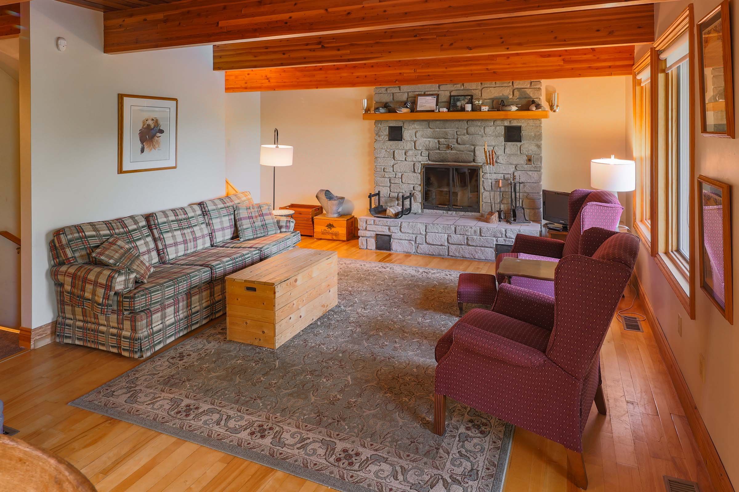 Living room with stone fireplace, wooden ceiling beams, plaid sofa, red armchairs, and a TV near windows.