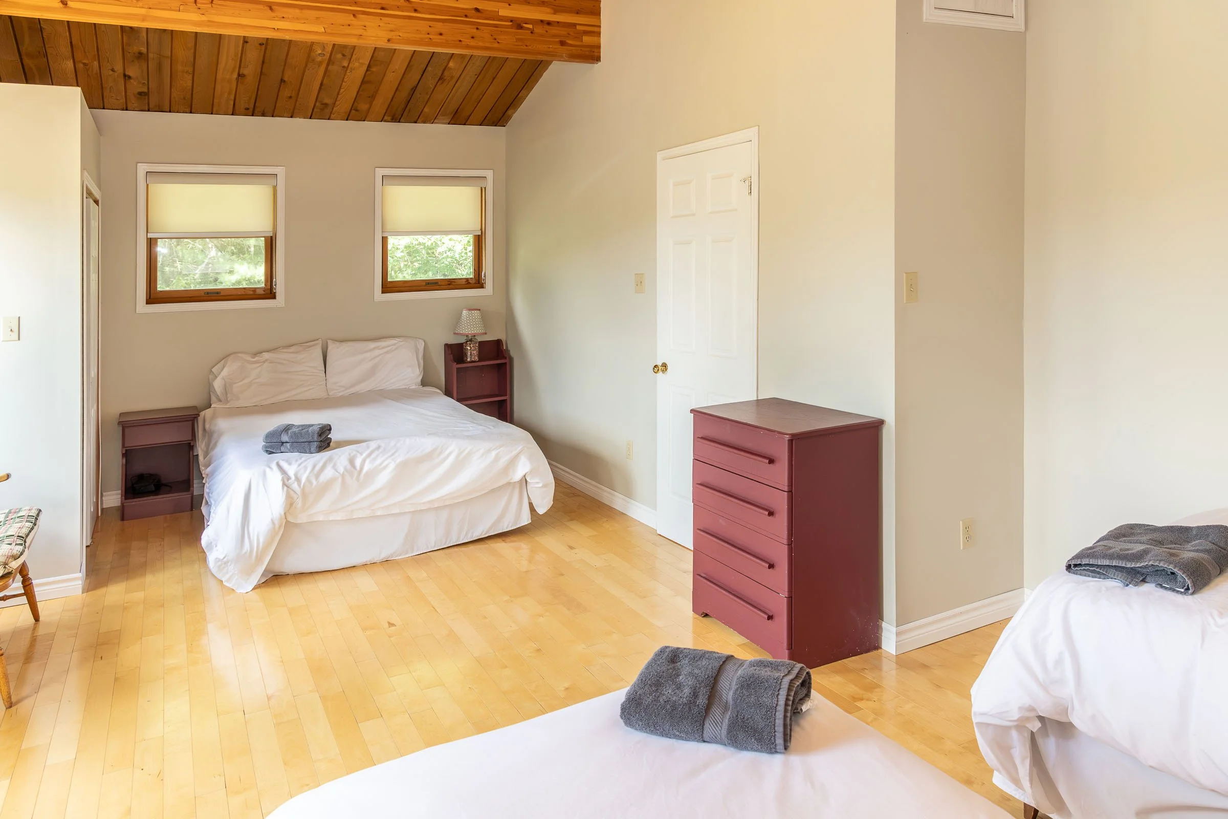 A bedroom with two beds, each with a gray towel on top. There are two small windows with yellow shades, a small lamp on a red nightstand, a white closet door, and a red chest of drawers. The room has hardwood floors and a wooden ceiling.