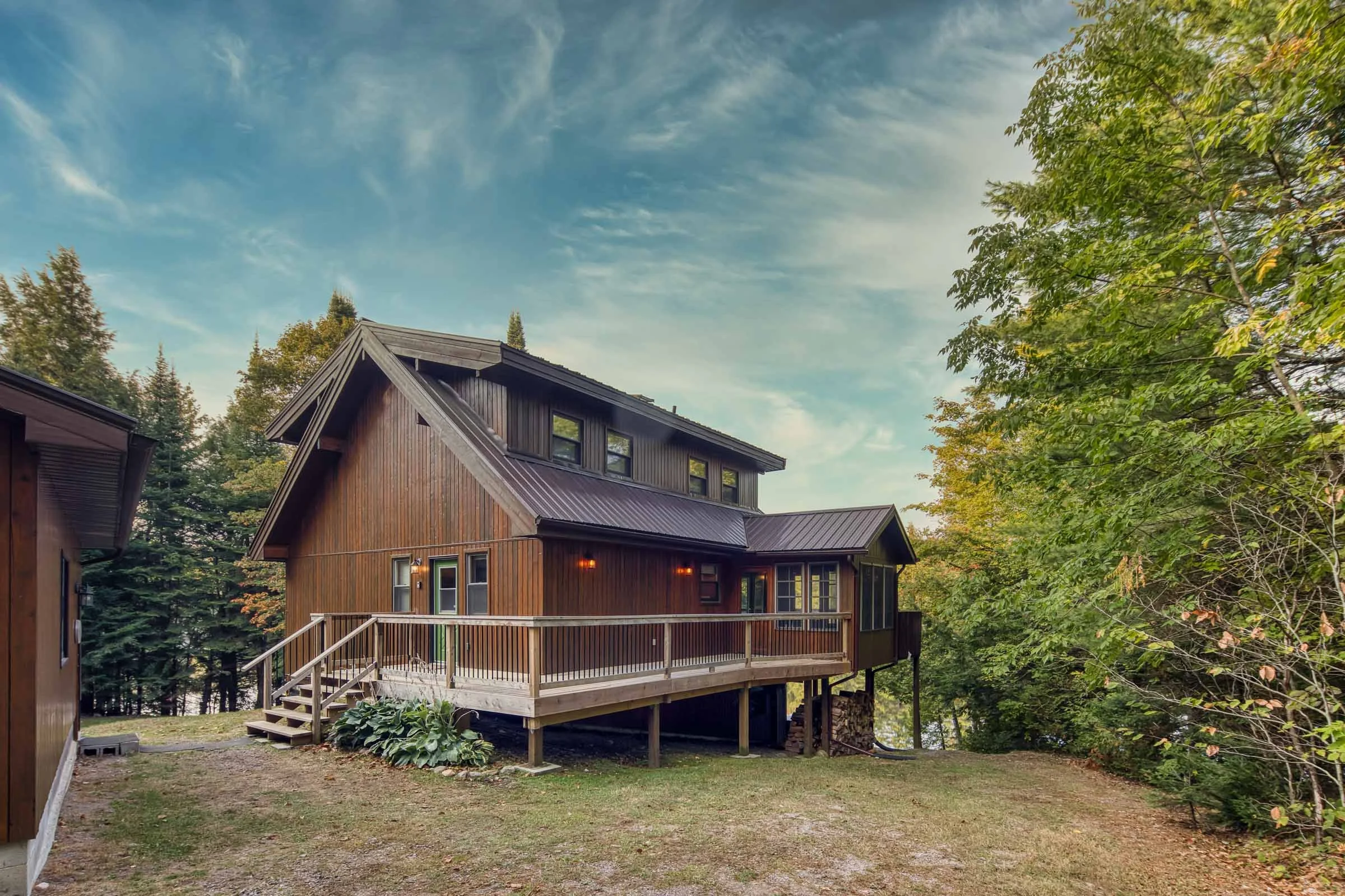 A wooden house with a deck, surrounded by trees with green leaves under a partly cloudy sky.
