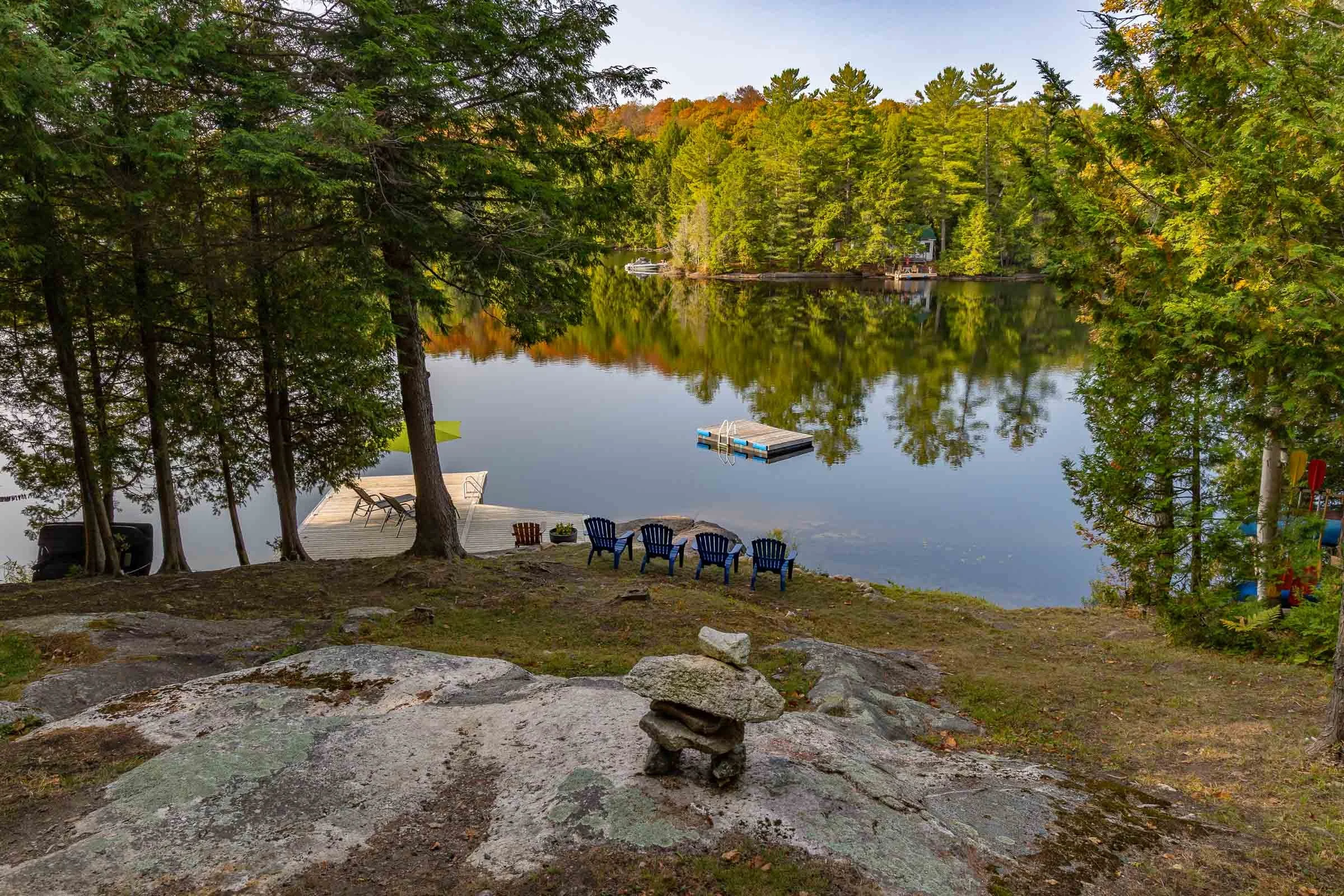 View of a calm lake surrounded by dense green trees with some autumn foliage, a dock with chairs on the shore, and a house reflecting in the water.