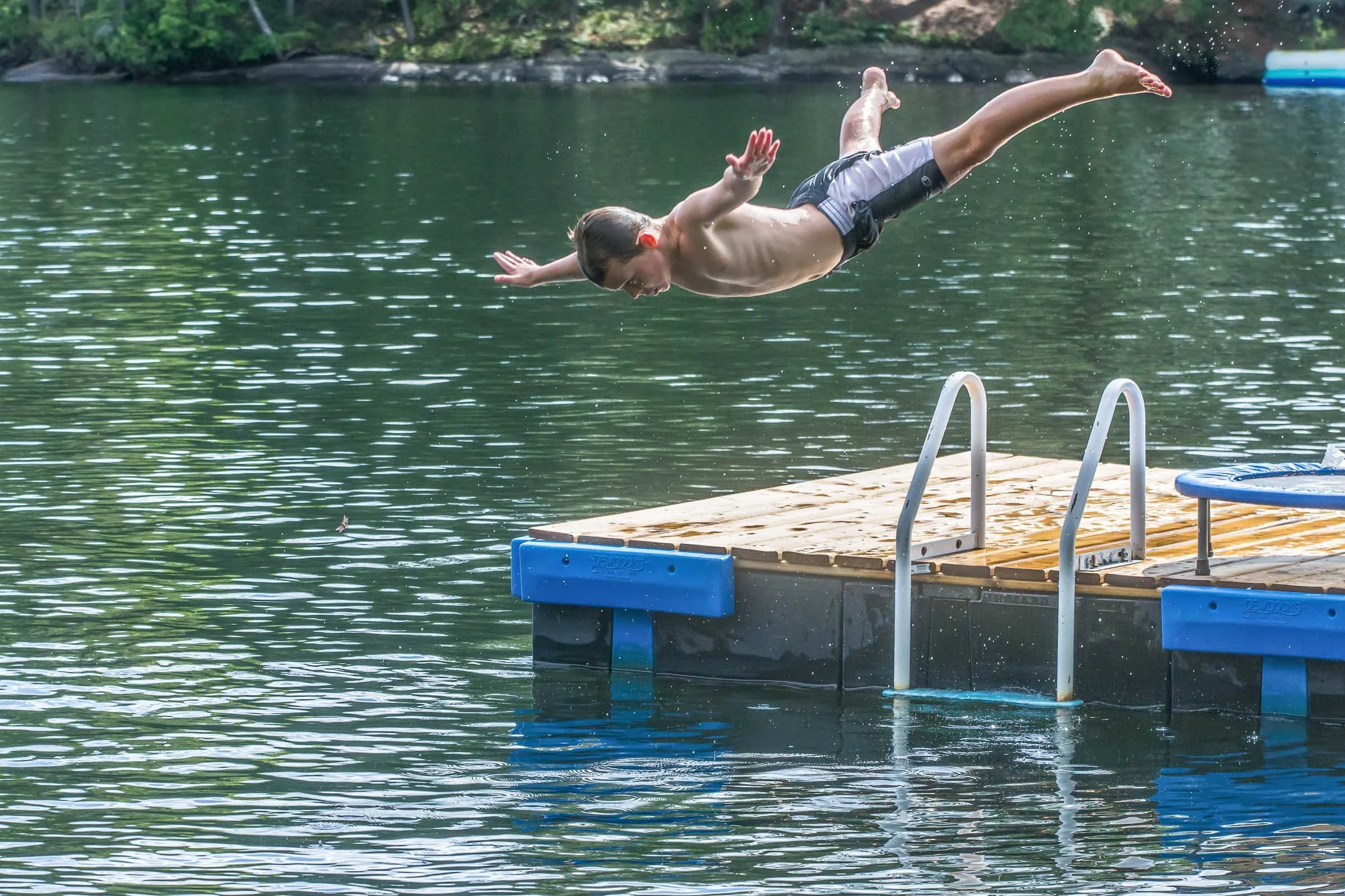 A young boy diving headfirst into a lake from a wooden dock surrounded by trees.