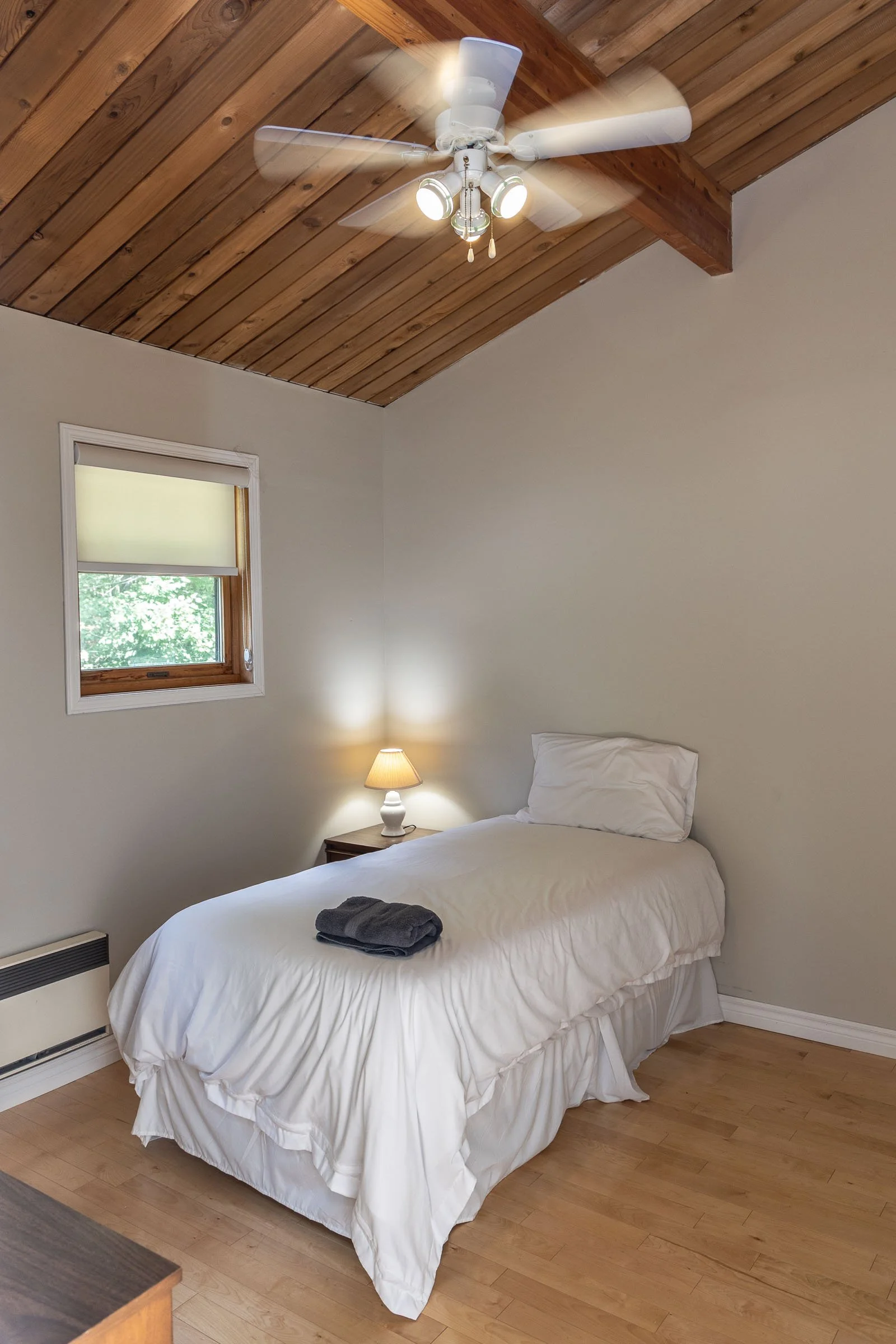 A bedroom with a bed covered in white bedding, with a folded towel on top. There is a small wooden nightstand with a lamp next to the bed. The room has a wood-paneled ceiling with a white ceiling fan and lights. There is a small window with a shade a