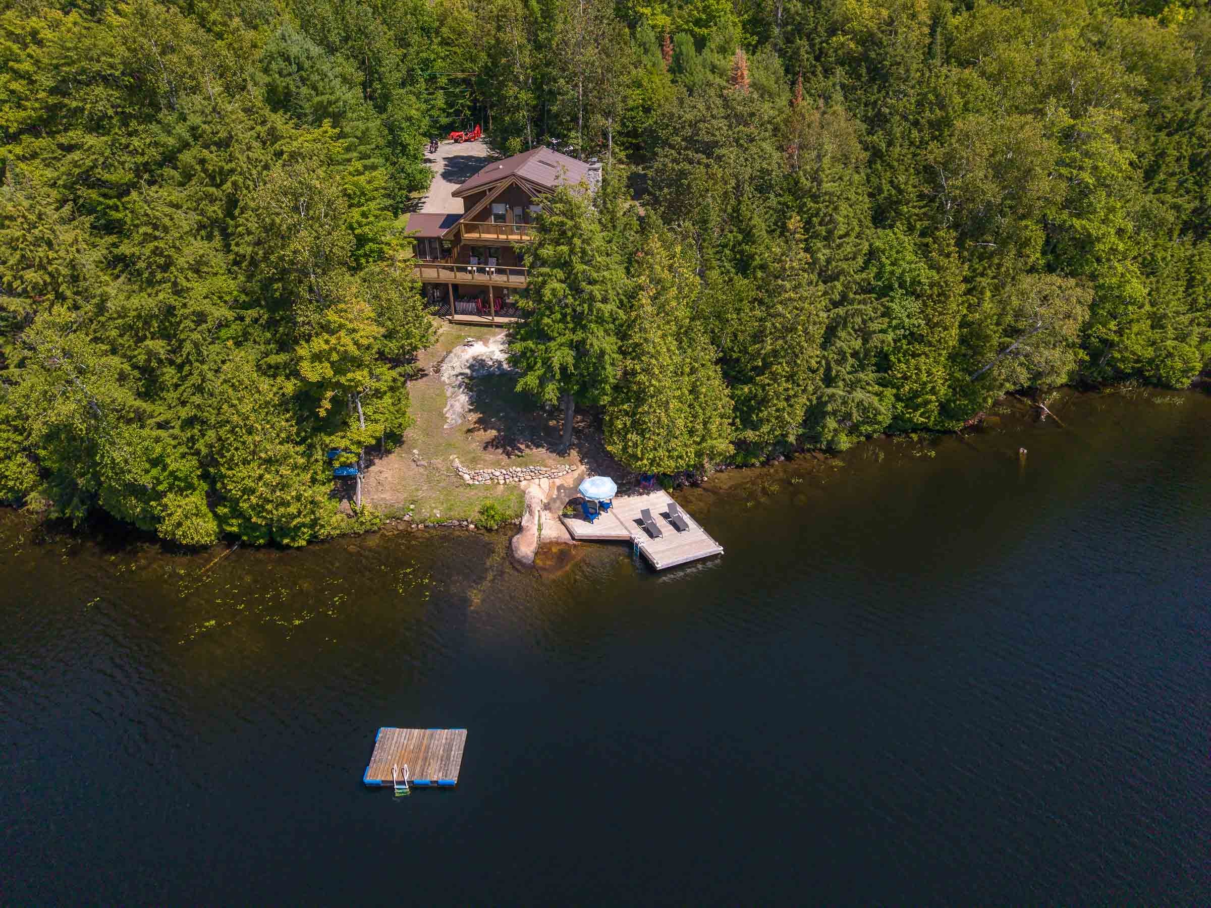 Aerial view of a lakeside house surrounded by dense green forest, with a small dock extending into the lake and a floating dock with a ladder on the water nearby.